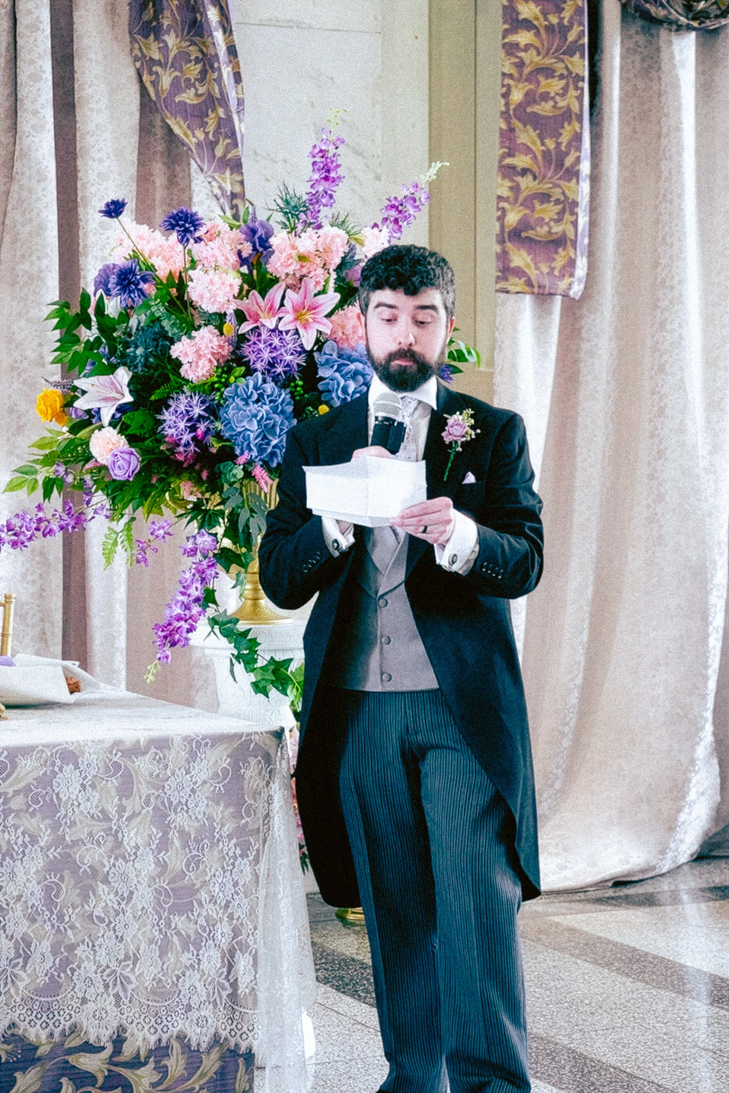 A man with dark curly hair and a beard dressed in a tuxedo with a boutonniere, reading from a piece of paper while standing next to a large pink, purple, and blue flower arrangement at a formal event or wedding.