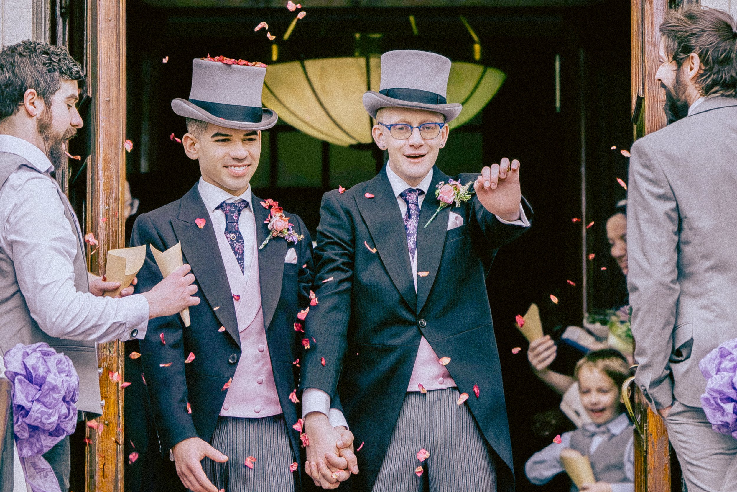 Two men holding hands and smiling, dressed in suits with gray top hats, at a wedding celebration with flower petals falling. A woman and children are visible in the background.