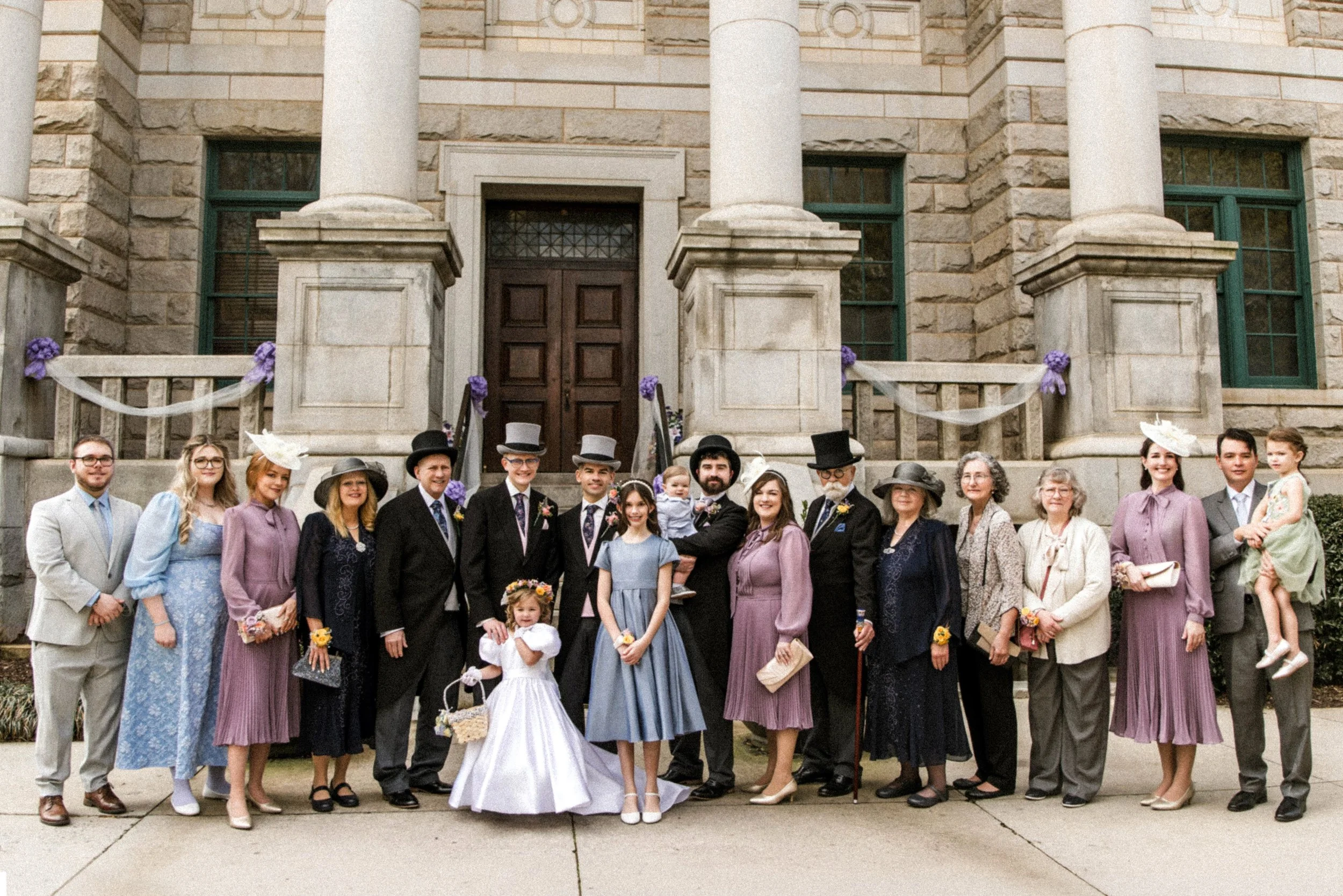 A large group of people dressed in vintage or historical clothing standing on the steps outside a stone building, possibly for a wedding or special event. Many are smiling and facing the camera.