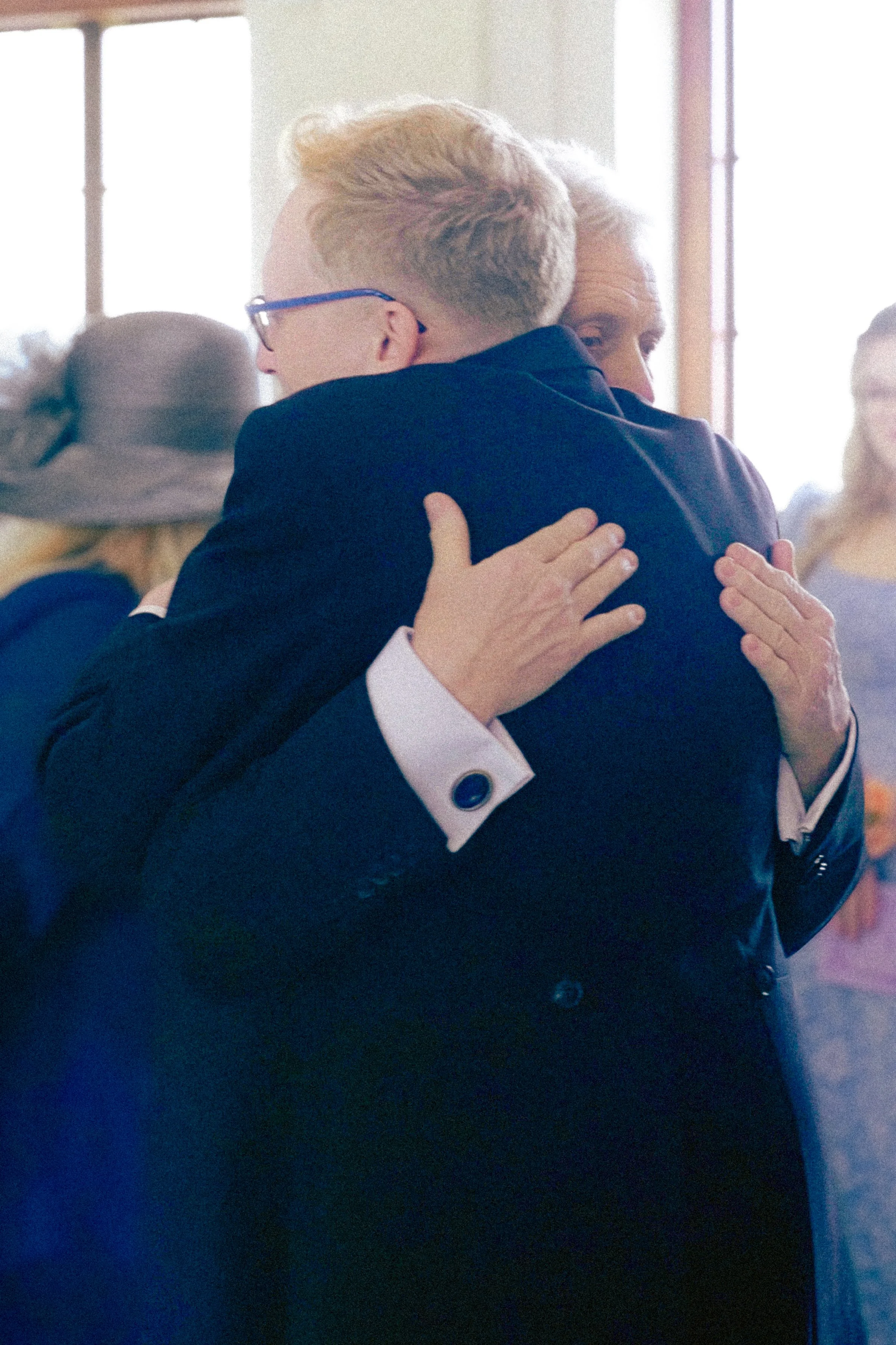 Two people hugging each other at what appears to be a social gathering or event. The person in the foreground has light blonde hair and is wearing glasses, while the person in the background has long dark hair and is smiling.