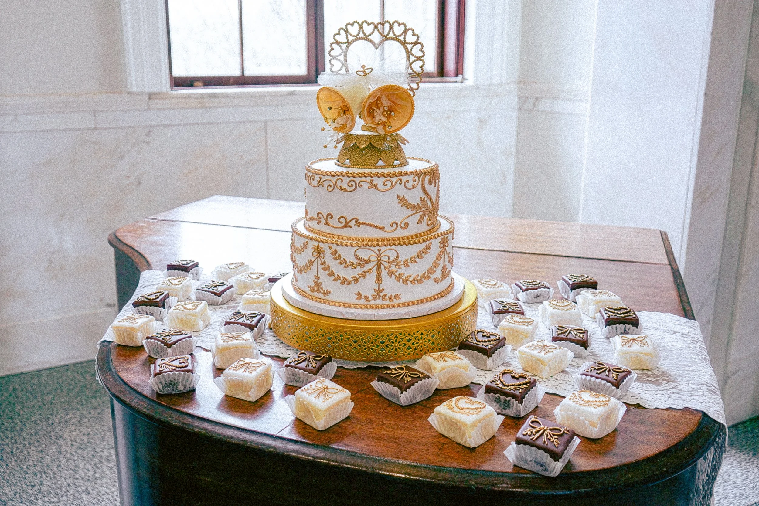 A three-tiered wedding cake with gold decorative piping and a heart-shaped topper featuring a butterfly with gold accents, surrounded by small decorative elements, is placed on a table with assorted small decorated desserts around it.