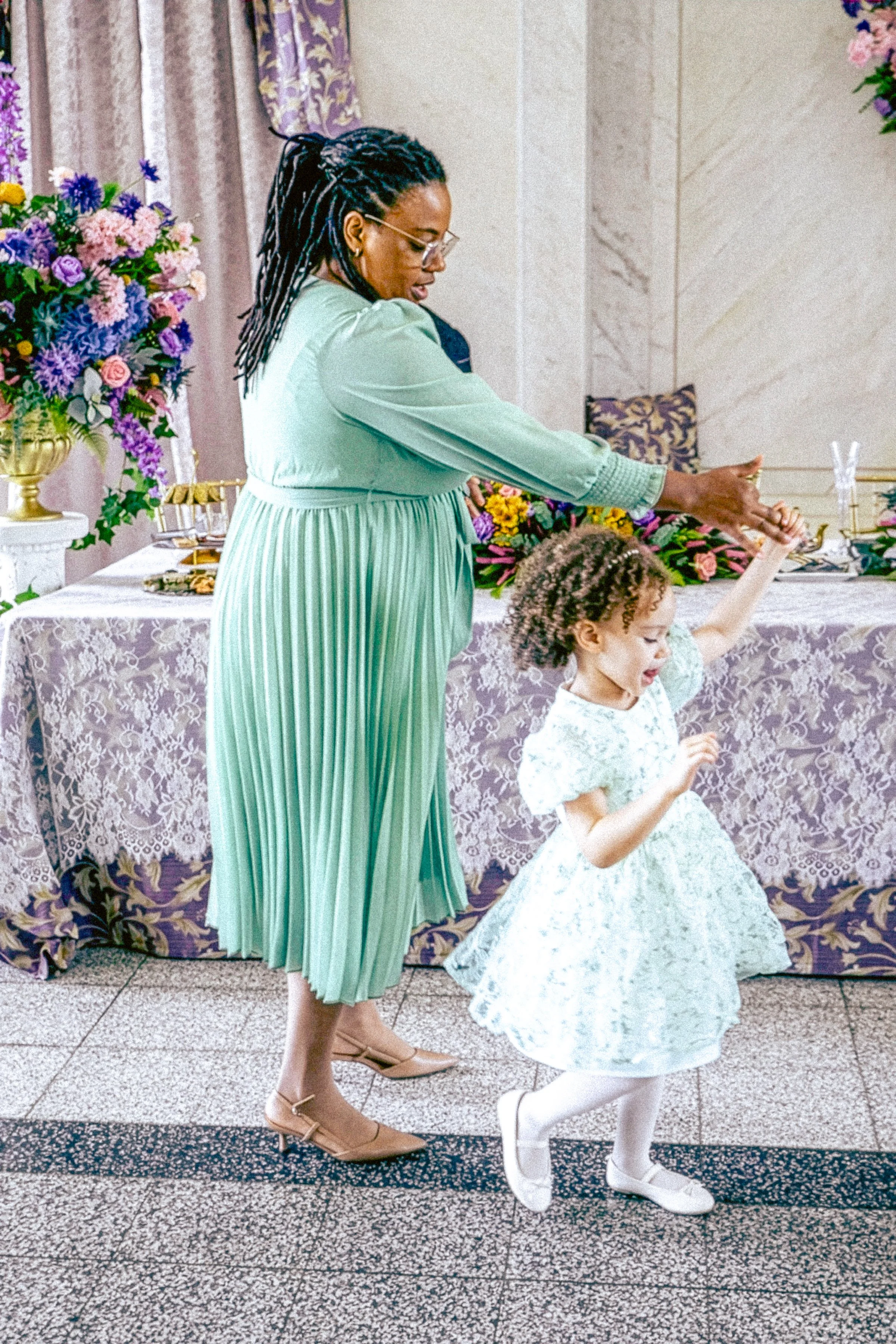 A woman with braided hair wearing a green dress is dancing with a young girl in a white floral dress at a party or celebration with floral arrangements and decorated table in the background.