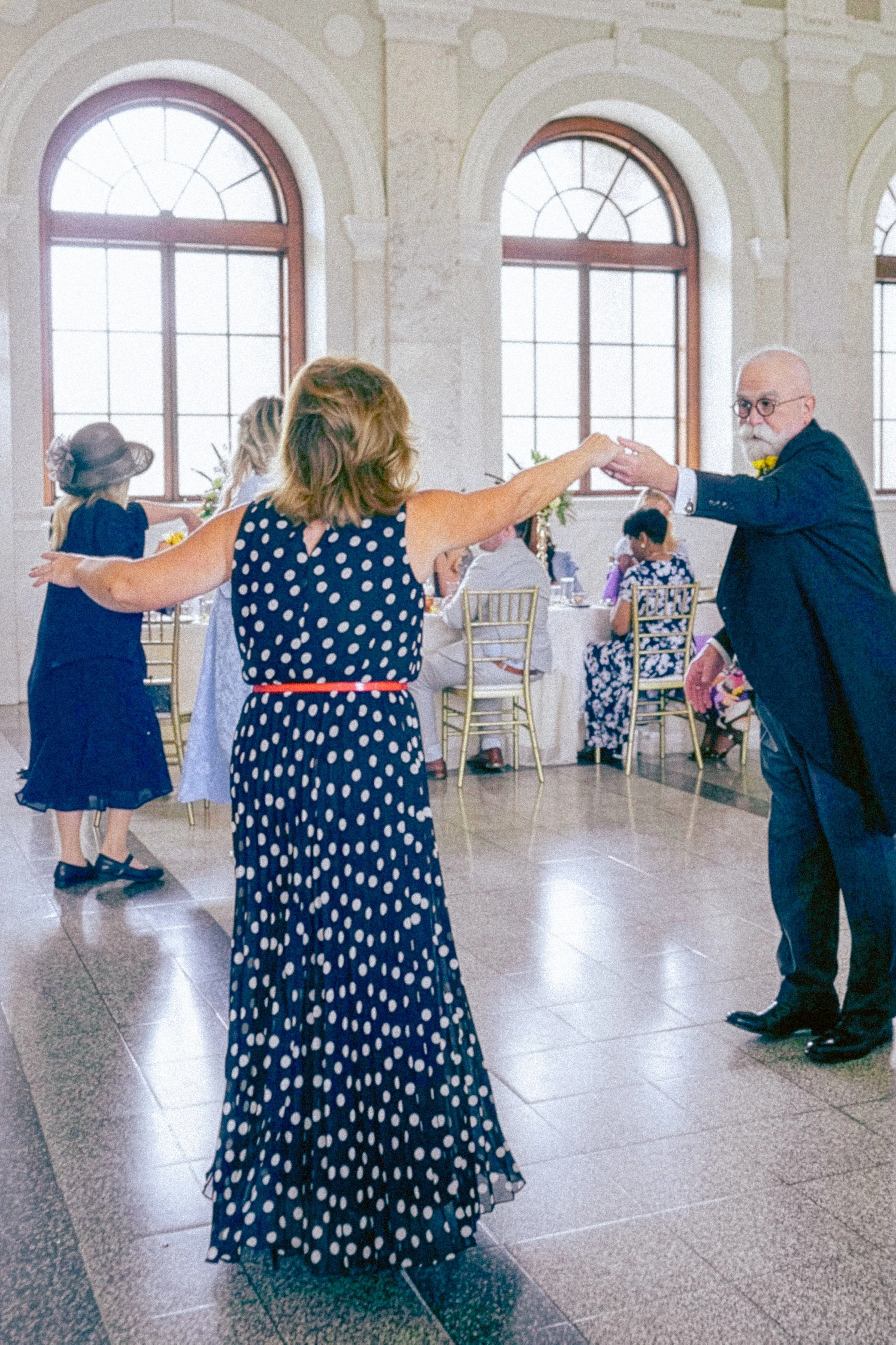 A woman in a polka dot dress dancing with a man in a suit at a social gathering in a room with large arched windows and tables with seated guests.