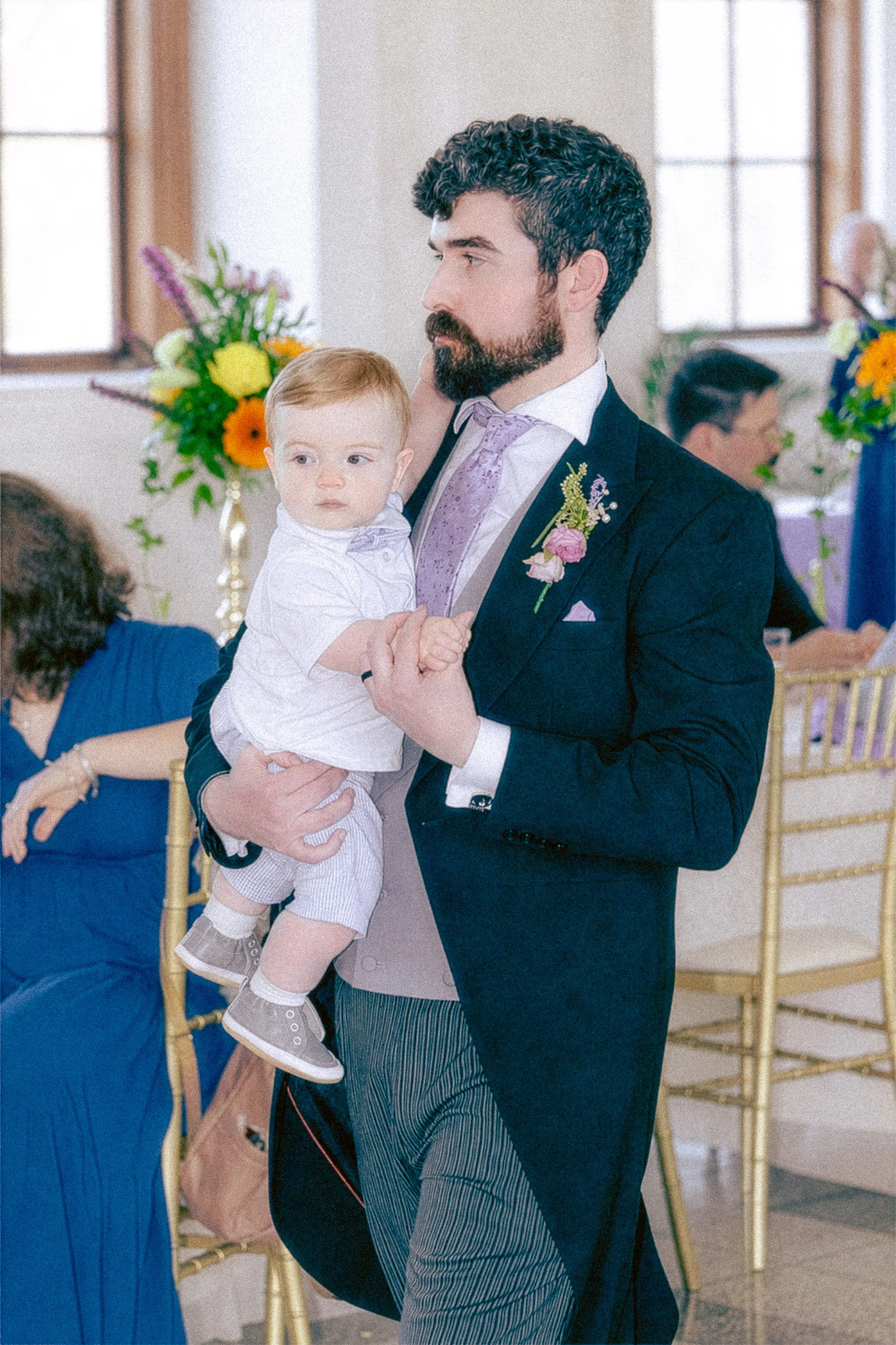 A man with curly dark hair and a beard dressed in a formal suit holding a young boy at a social event. The man is wearing a navy blazer with a flower boutonniere, a lavender tie, and a white shirt. The boy has light brown hair, wears a white shirt, b