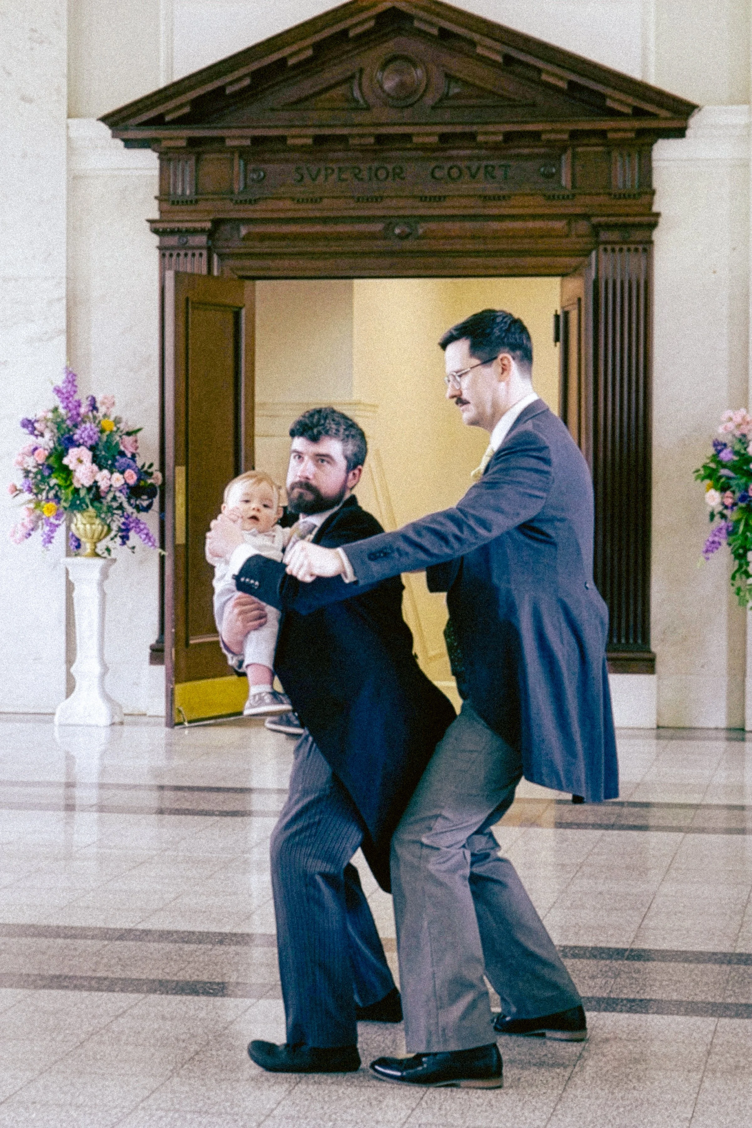 Two men in suits are dancing in a corridor, with one holding a young child. The corridor has floral arrangements on stands and a wooden archway inscribed with 'Superior Court' above the doorway.