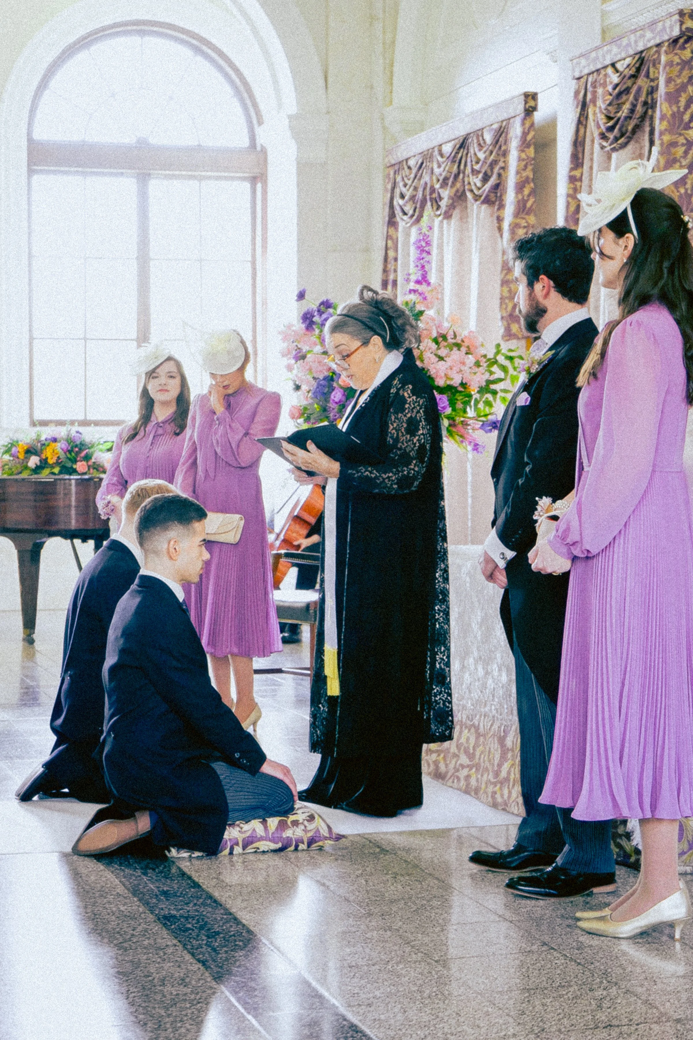 A couple kneels in front of a woman reading from a book during a wedding ceremony inside a grand hall with tall windows and ornate curtains. The woman officiating wears black lace, and the bride and groom are dressed in formal attire, with the bride 