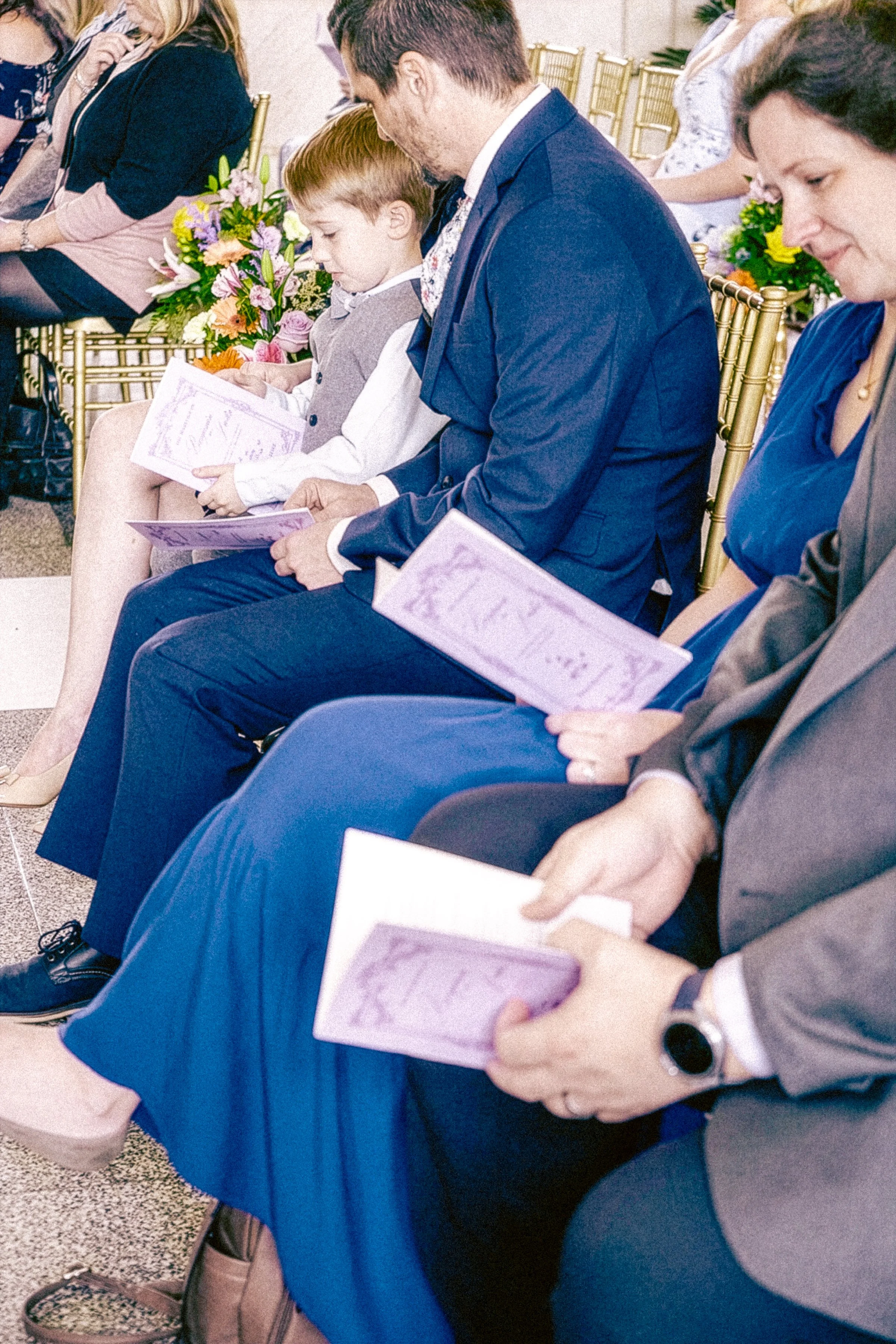 People seated in chairs at an event, holding brochures or programs, with flower arrangements visible in the background.