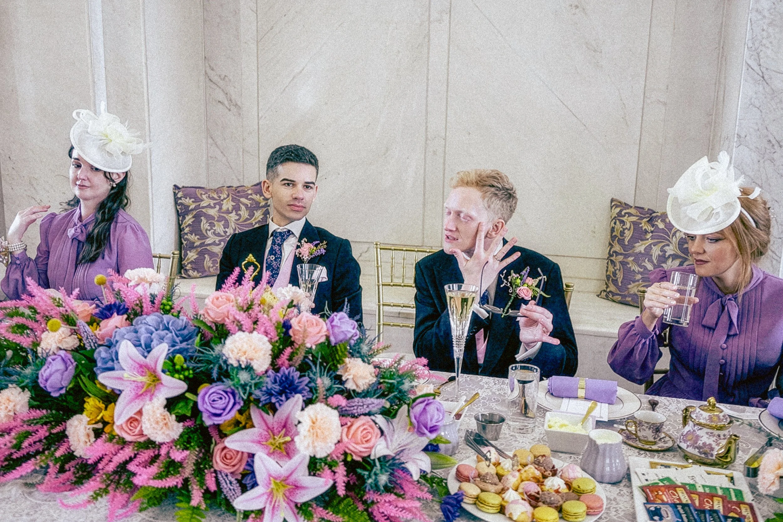 People seated at a formal event table decorated with a large colorful flower arrangement, with several women in purple dresses and white hats, and men in suits, engaging in conversation, with food and drinks on the table.