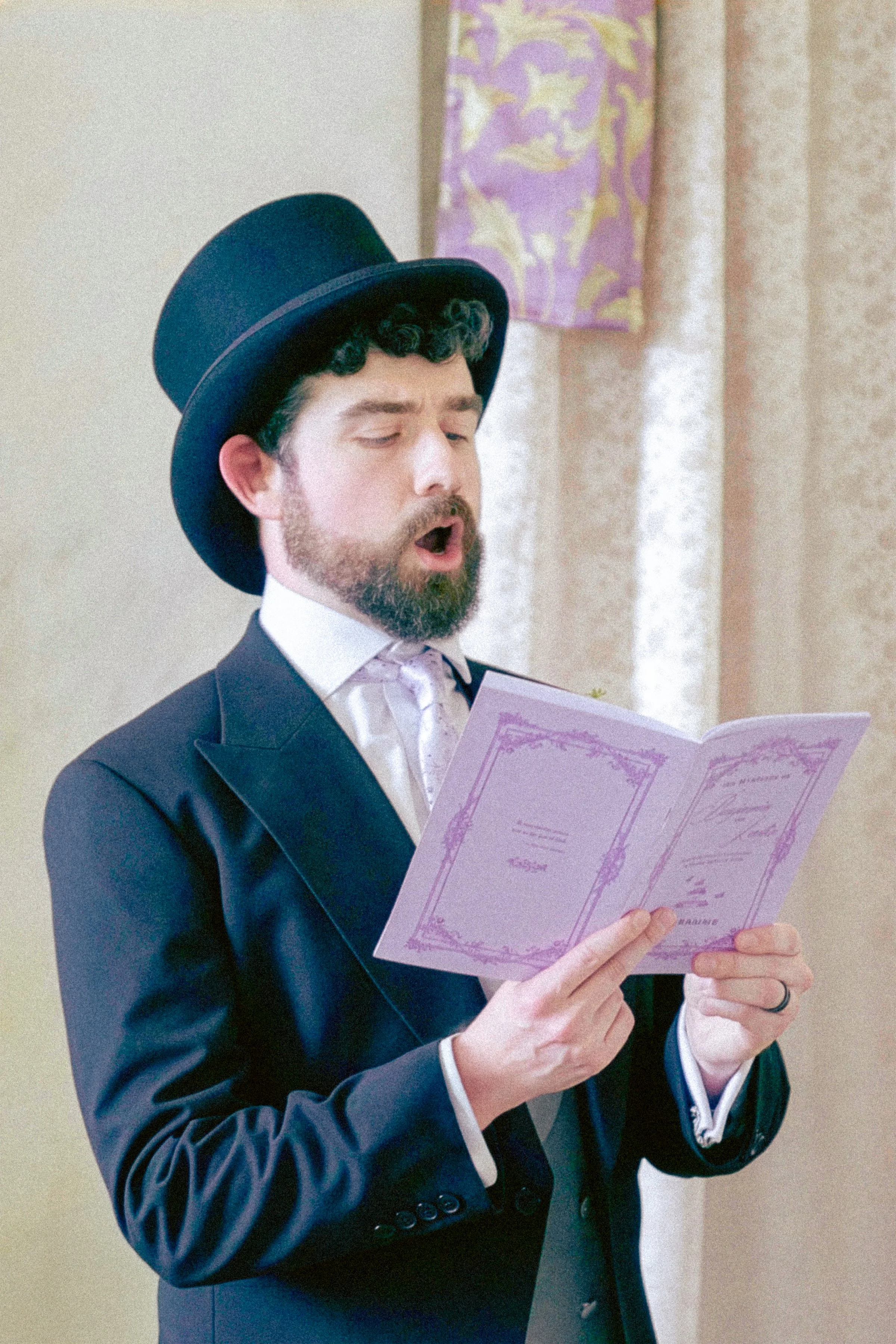 A man in a formal dark suit with a vest, white shirt, and striped tie, wearing a large black top hat and holding a pink paper with decorative borders and text, appears to be singing or reading aloud with his eyes closed.