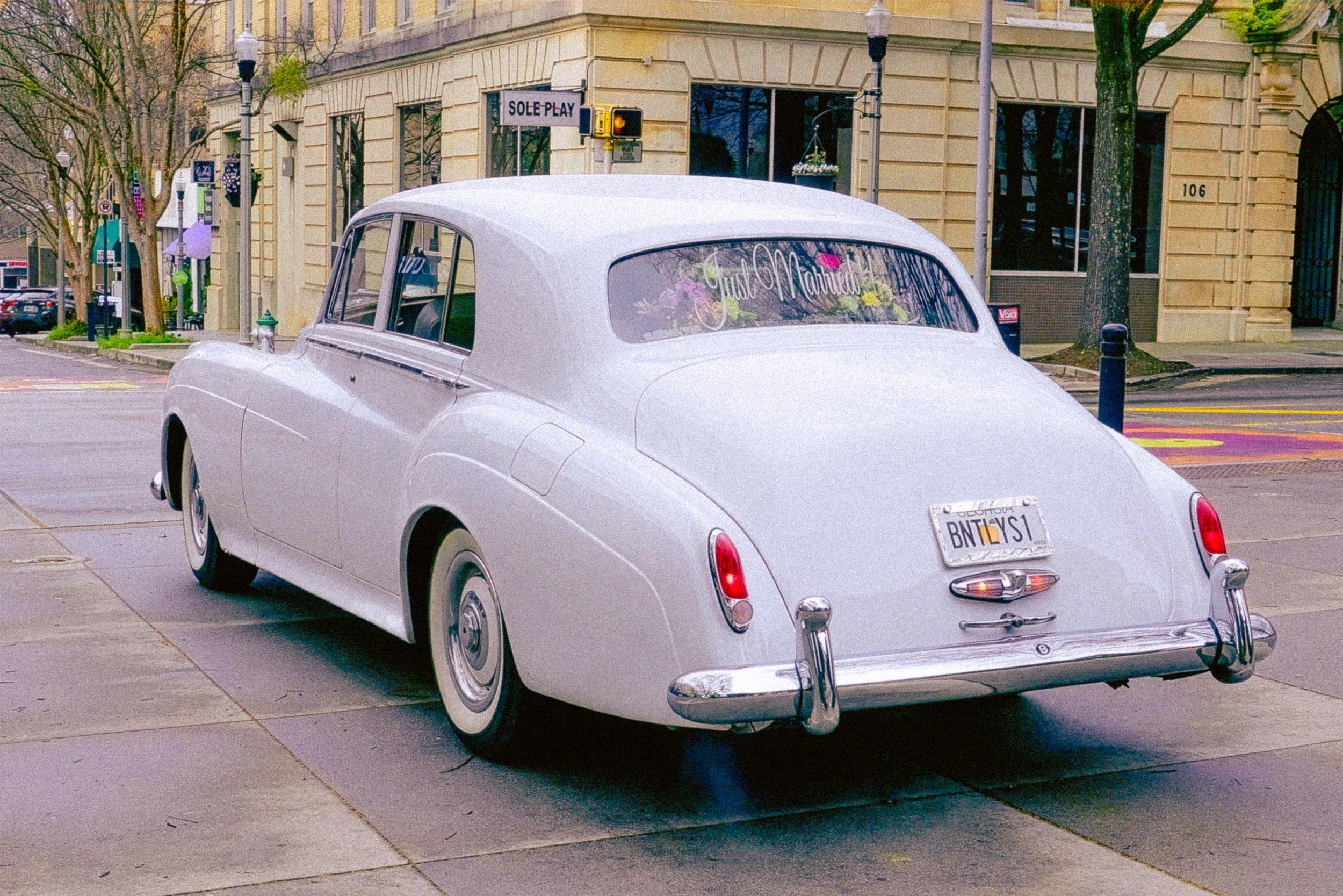 A vintage white car parked on a city street with a rear window decal that reads 'Just Married' with flowers inside the car. There are buildings, trees, and street signs in the background.