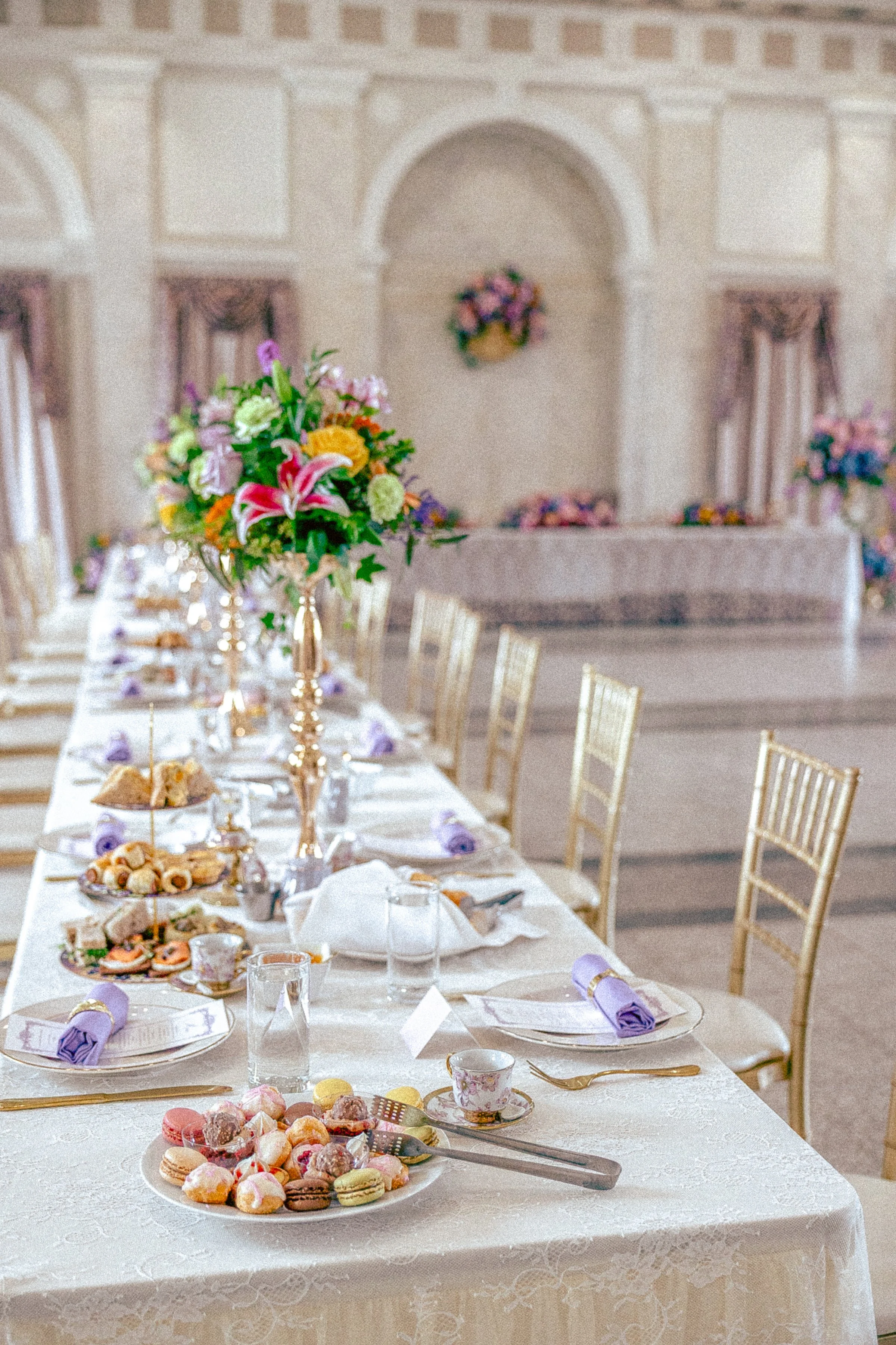 A banquet hall decorated for a celebration with a long table set with floral centerpieces, plates of assorted pastries and cookies, and purple napkins. There are gold chairs around the table, and the background features ornate decor and draped window