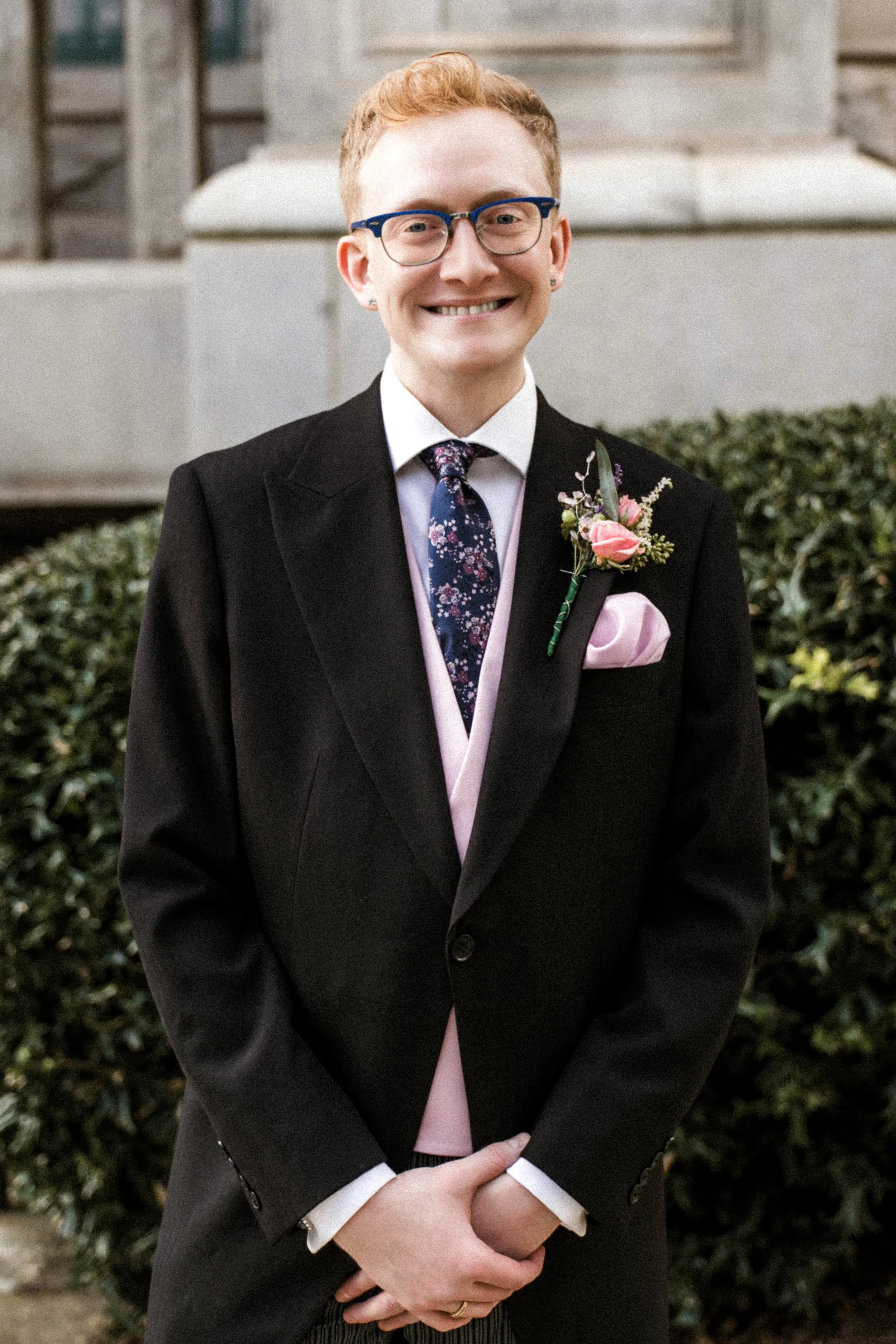A person in a black suit with a floral tie and pink bow tie, with a pink boutonniere, smiling outdoors in front of bushes and a building.