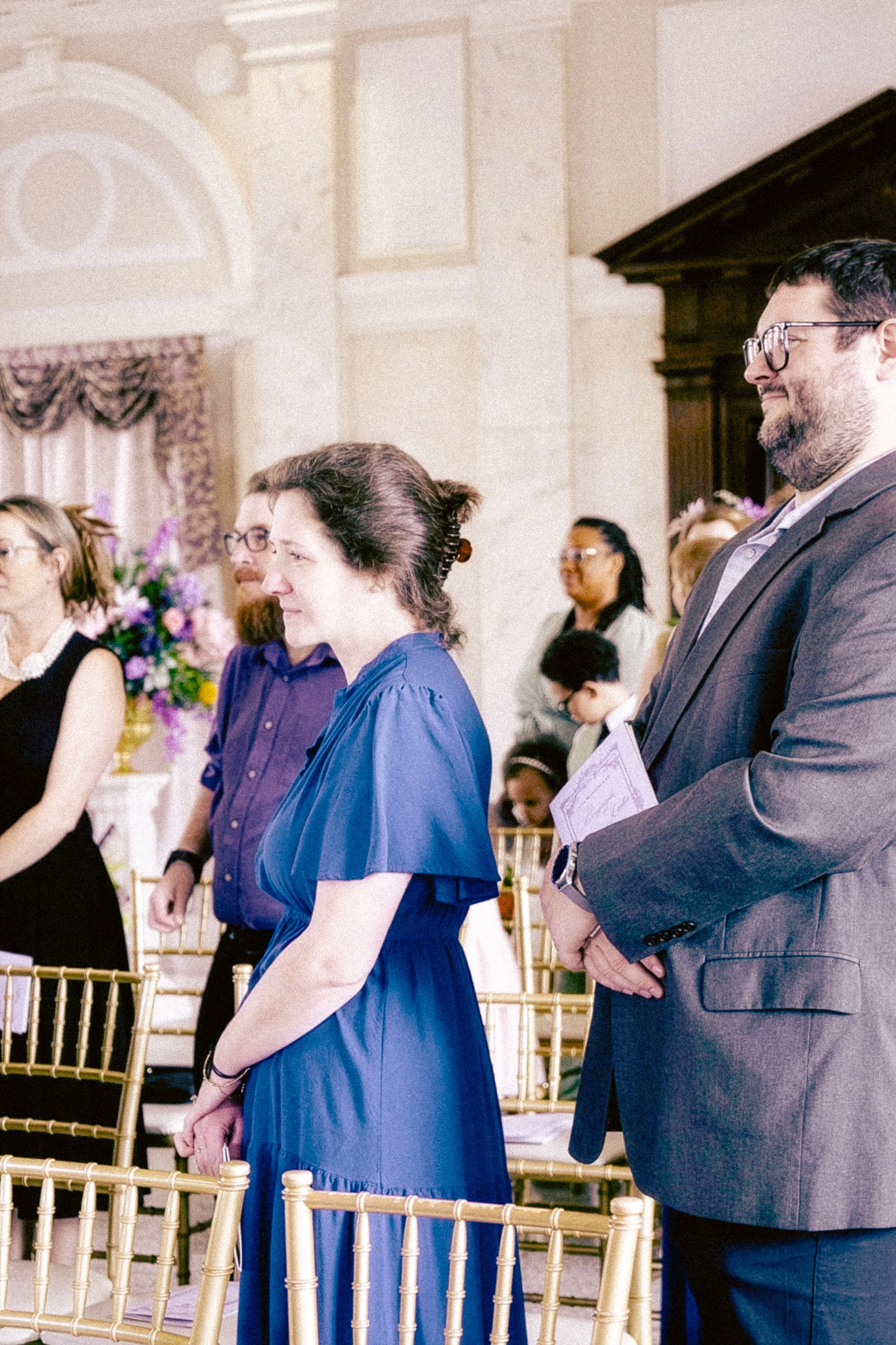 A group of people standing in a formal setting, with one man in a suit and a woman in a blue dress in the foreground, all appearing to participate in an event or ceremony.