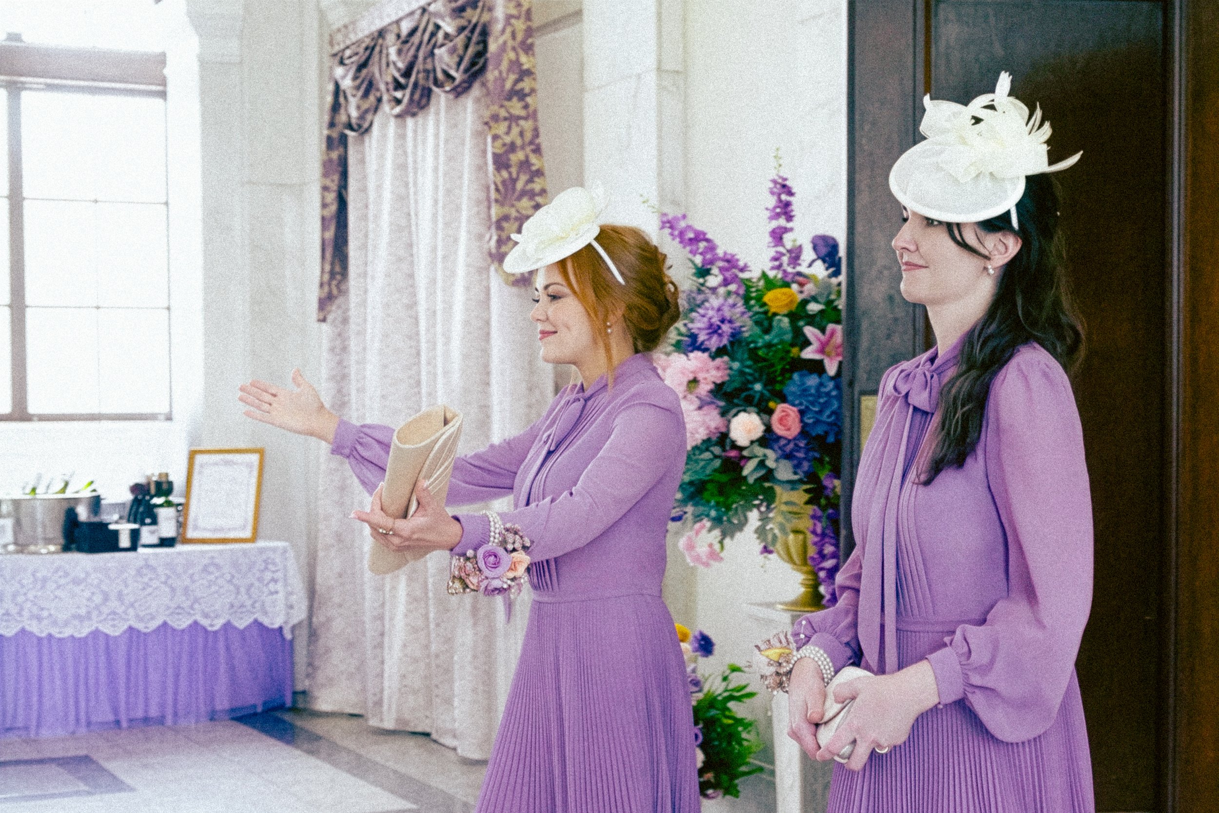 Two women in purple dresses wearing white hats with floral decorations at a formal event with floral arrangements and a decorated table in the background.
