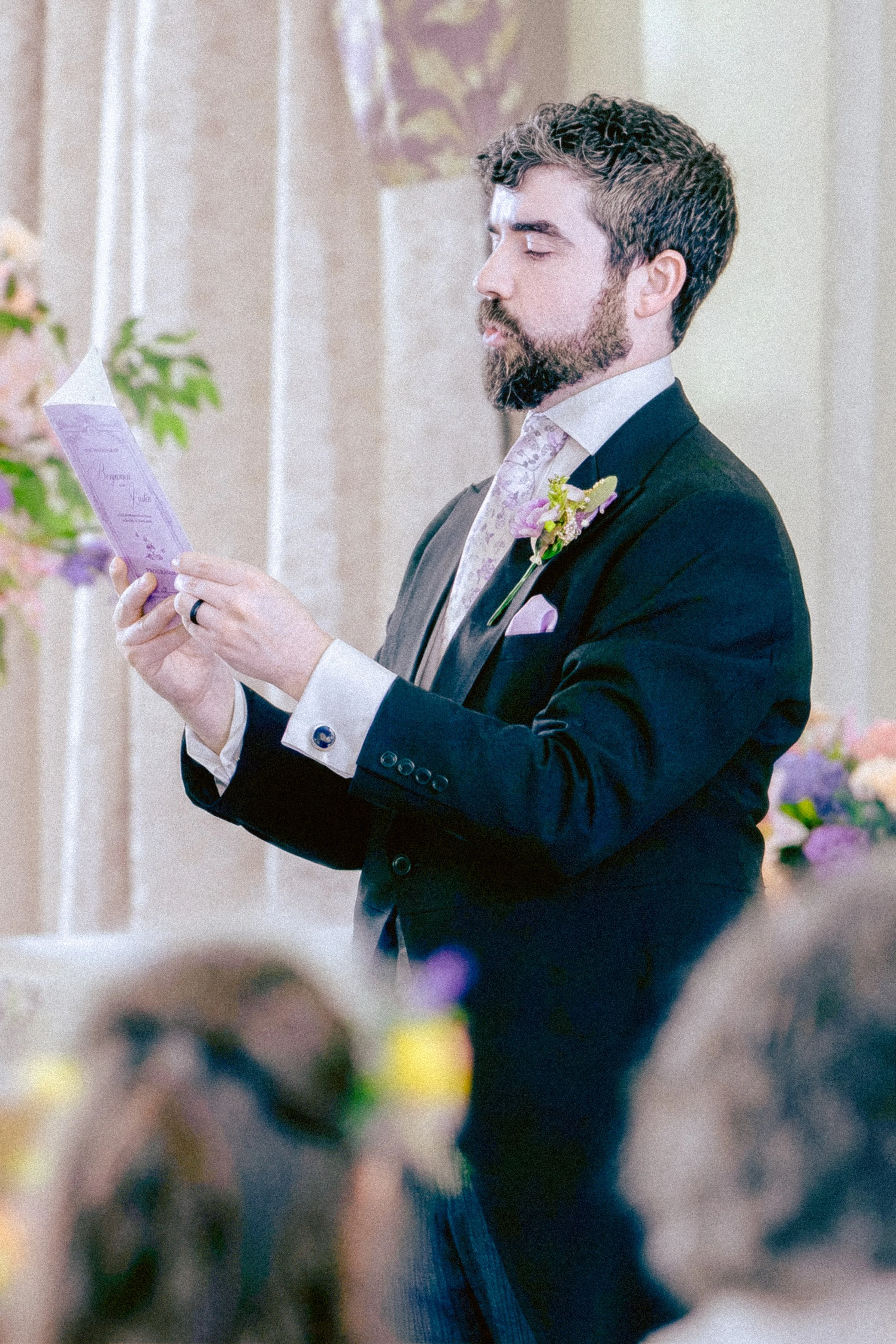 A man with a beard and styled hair, dressed in a dark suit with a light-colored shirt and floral tie, stands indoors during a wedding ceremony, reading from a piece of paper.