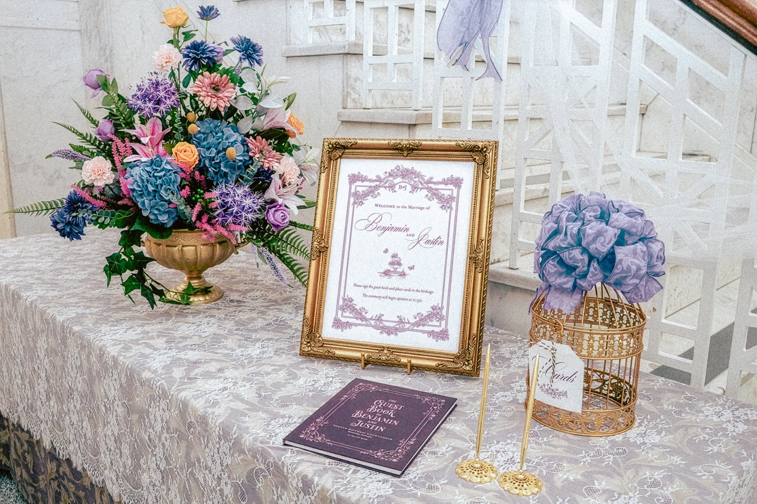 A wedding reception table with a large colorful floral arrangement in a gold vase, a framed wedding invitation for Benjamin and Justin, two gold decorative candle holders, a guest book, and purple tulle balls in a gold wire stand, set against a beige