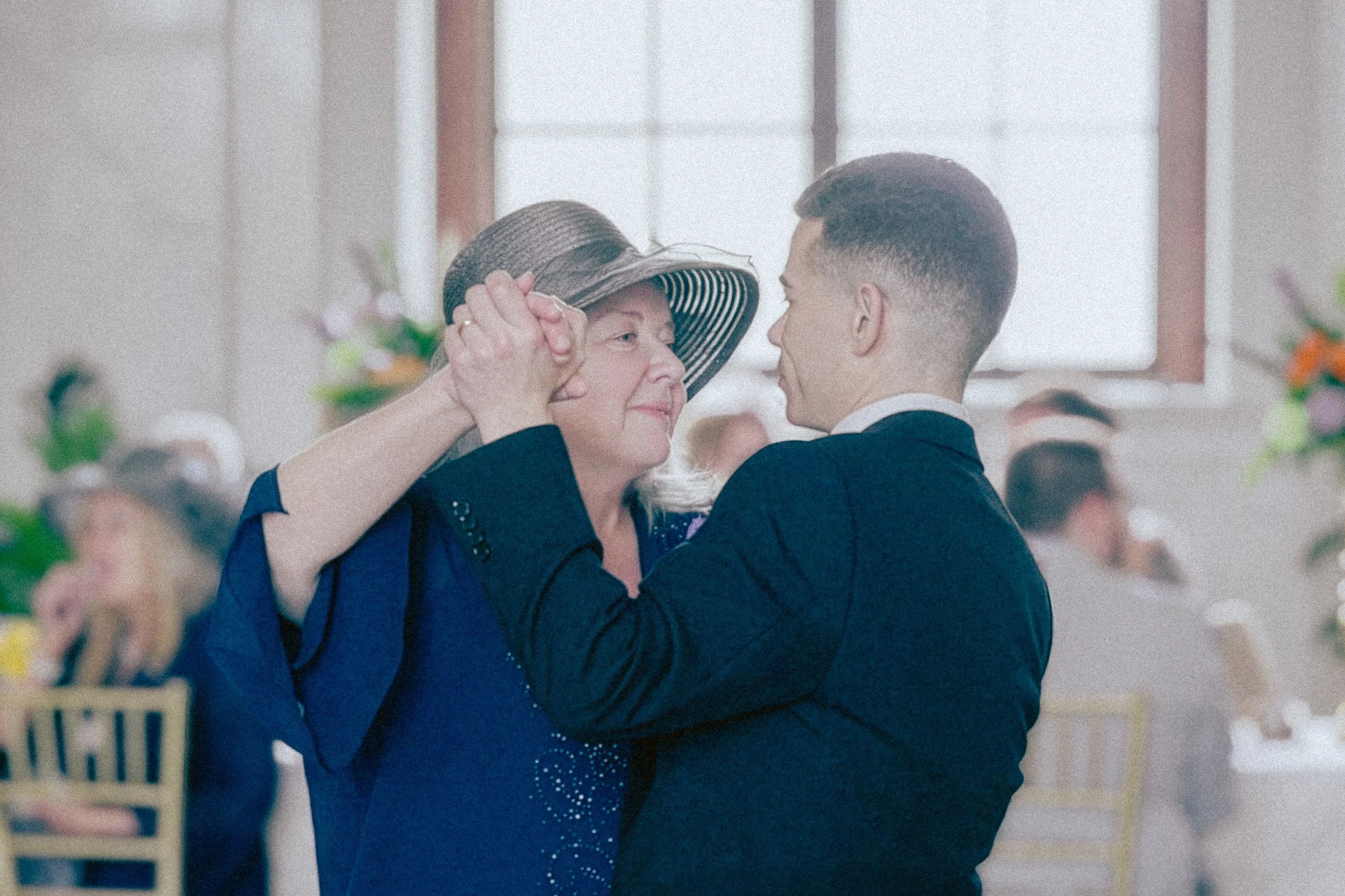An elderly woman and a young man are dancing close together, touching foreheads, with the woman's hand on the man's shoulder and the man gently holding her head. They are indoors in a brightly lit room with a large window in the background.