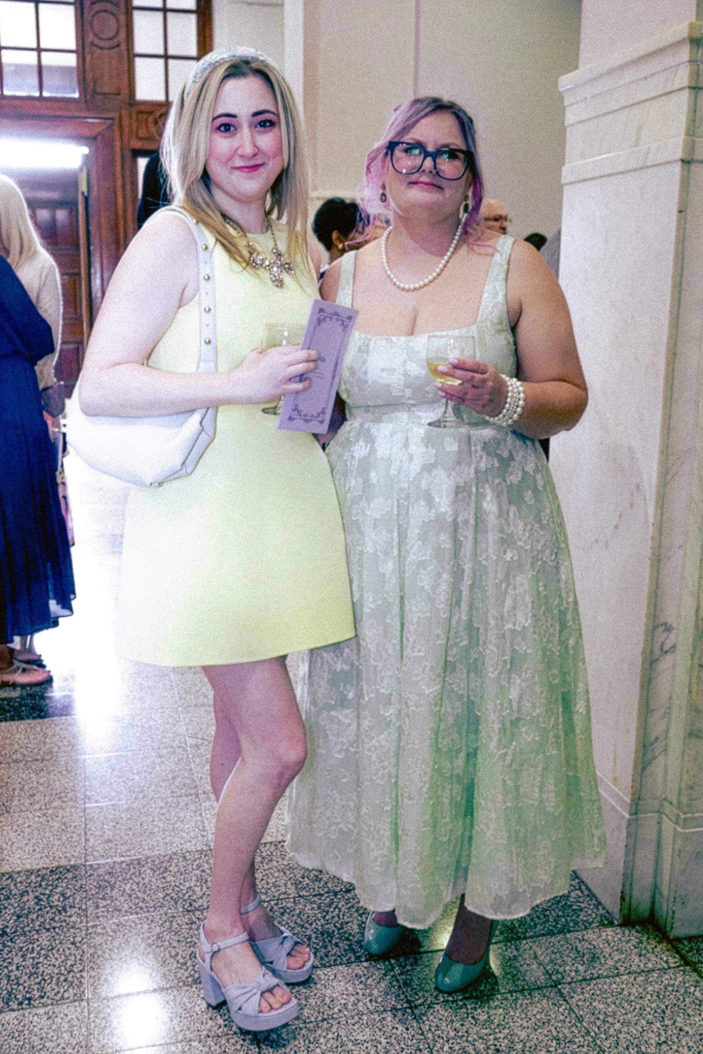 Two women at a formal event, dressed in vintage-style dresses. One woman is blonde with glasses and purple hair, holding a glass of white wine. The other woman has blonde hair and is wearing a yellow dress, holding a purple piece of paper. They are s