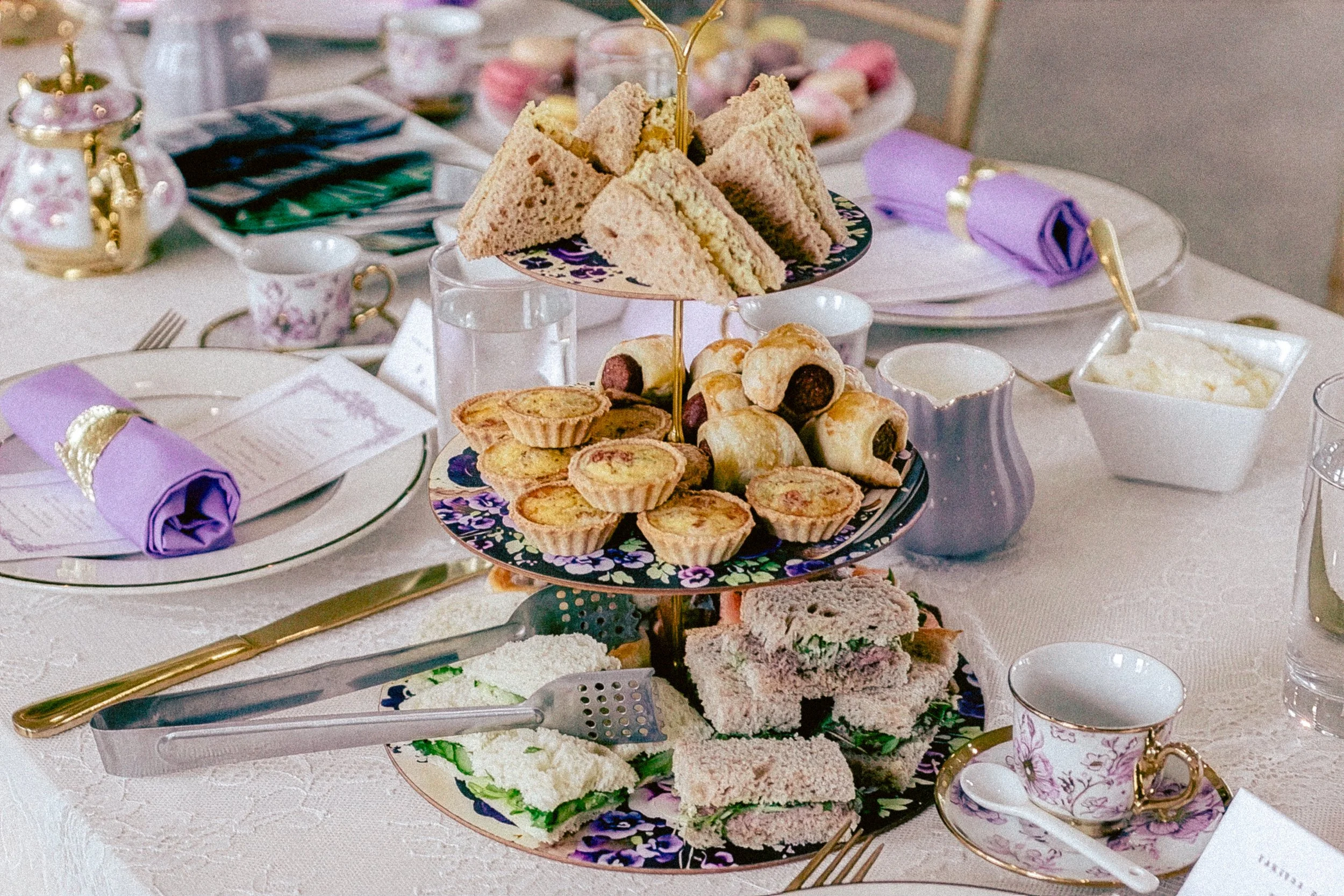 A multi-tiered tray filled with assorted sandwiches, pastries, and finger foods at an elegant tea party setting. The table is set with floral china, purple napkins, and gold utensils.