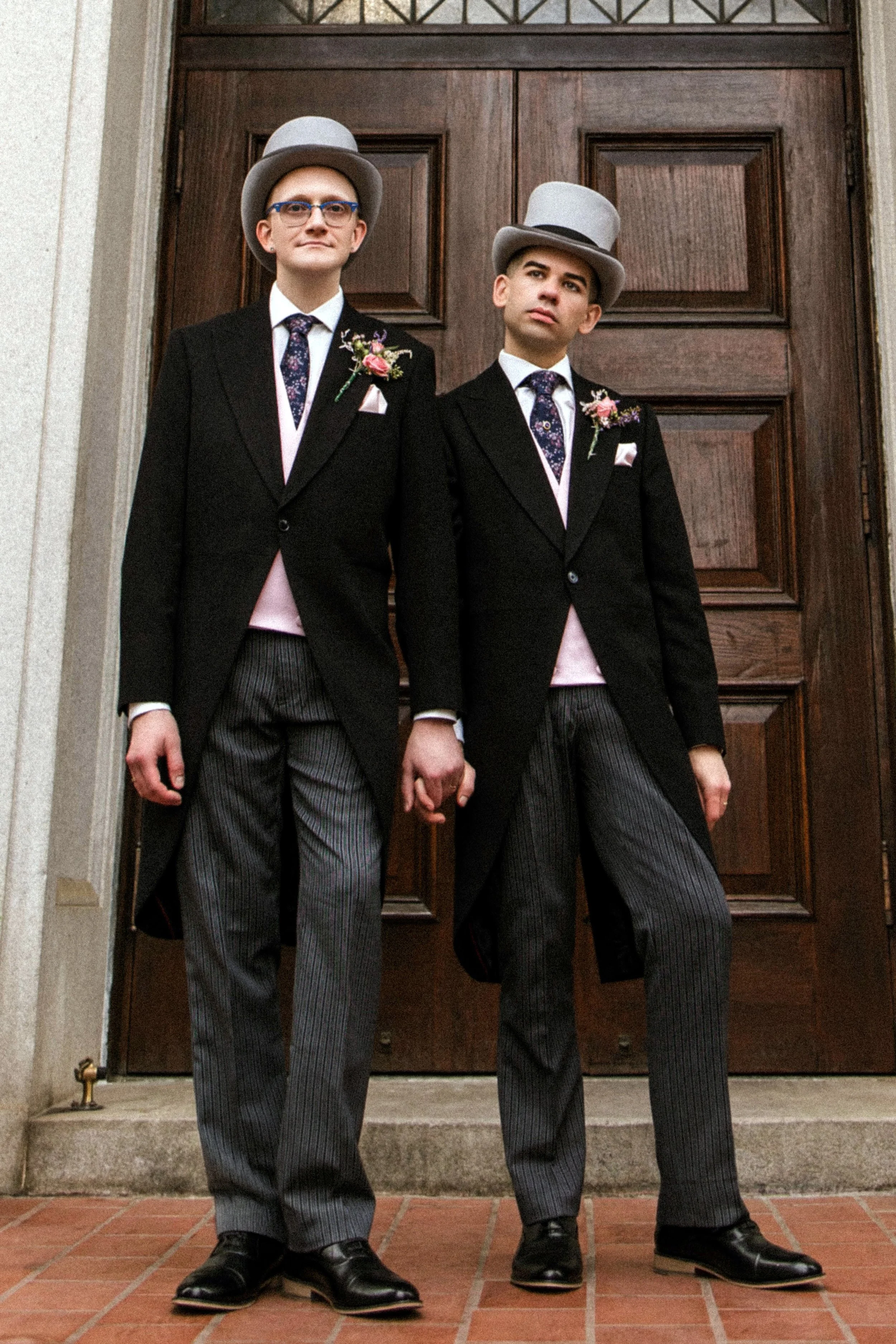 Two young men dressed in black tuxedos with pink vests and floral boutonnieres, wearing gray top hats and glasses, standing on steps in front of a wooden door.