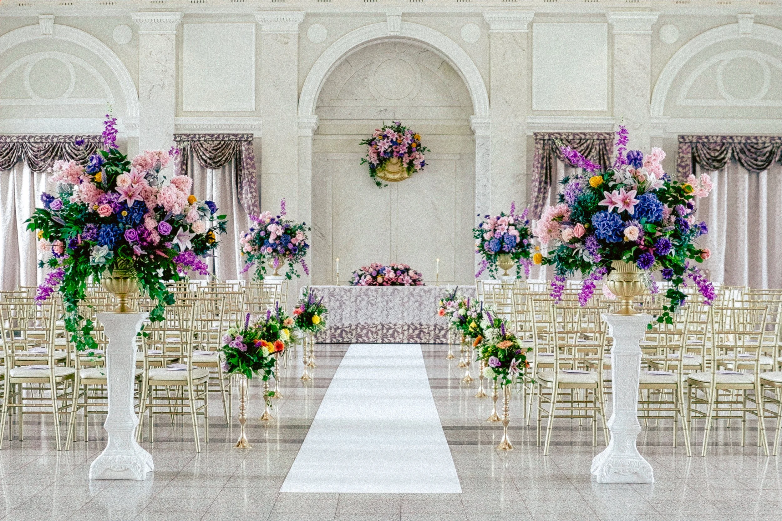 Elegant wedding altar decorated with large colorful floral arrangements and a white aisle runner in a grand hall with ornate architectural details.