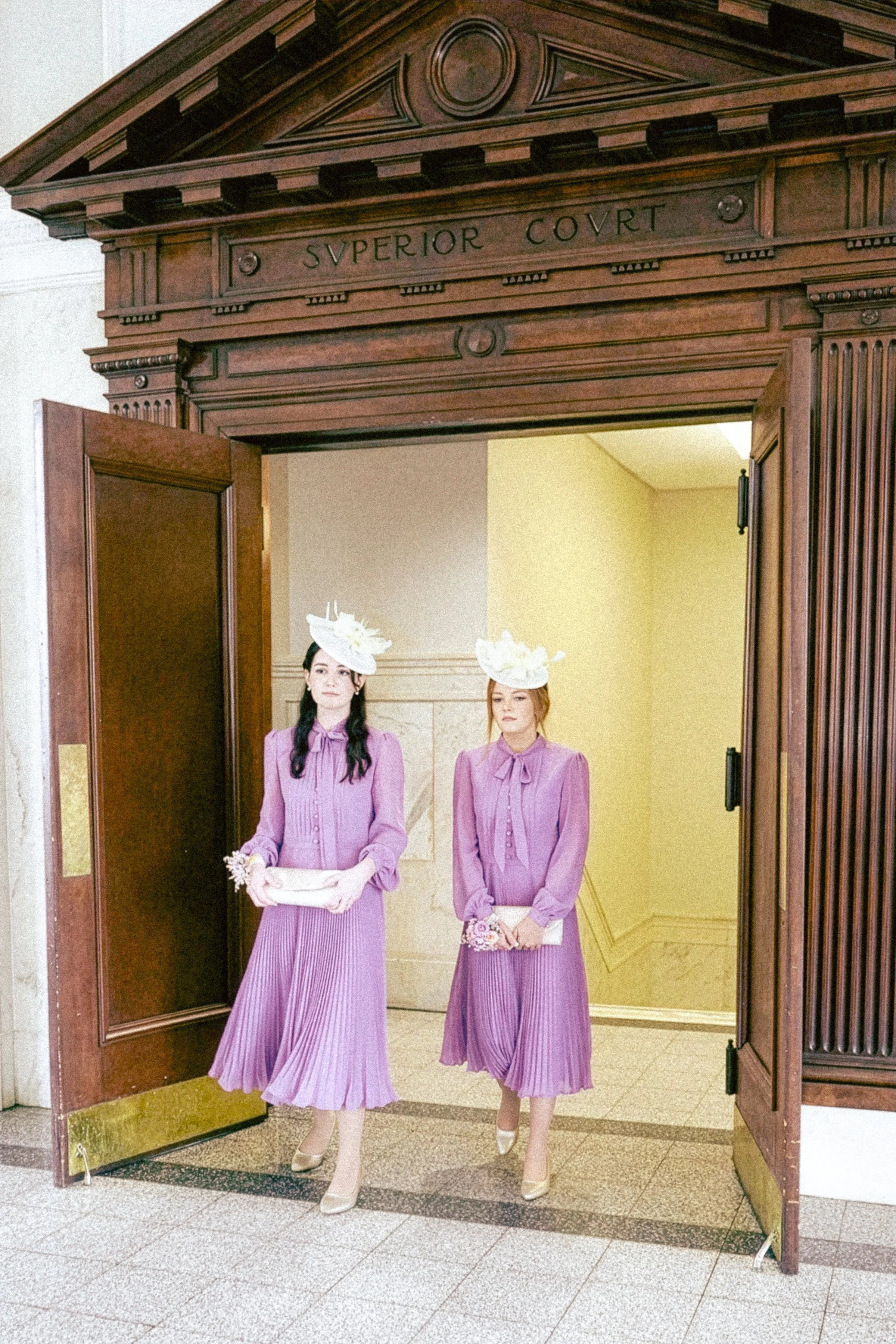 Two women in vintage purple dresses with hats and gloves standing in front of a wood-paneled doorway inside a building.
