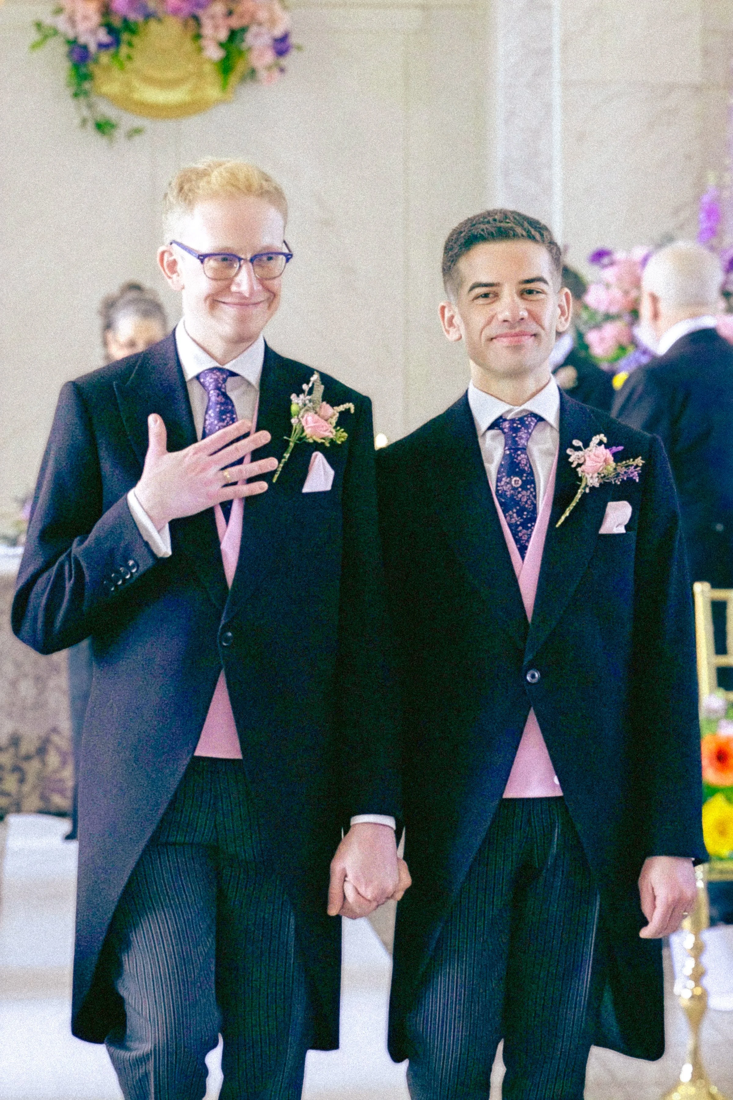Two men dressed in formal suits with boutonnières on the lapels, standing at a wedding ceremony, holding hands and smiling.