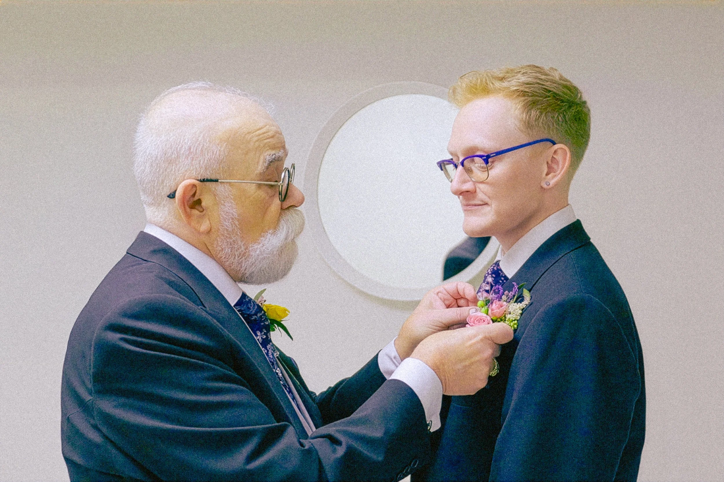 An elderly man with white hair and beard, wearing glasses and a dark suit, is pinning a pink boutonniere on a young man with short red hair and glasses, who is also dressed in a dark suit. They are facing each other and smiling, in what appears to be