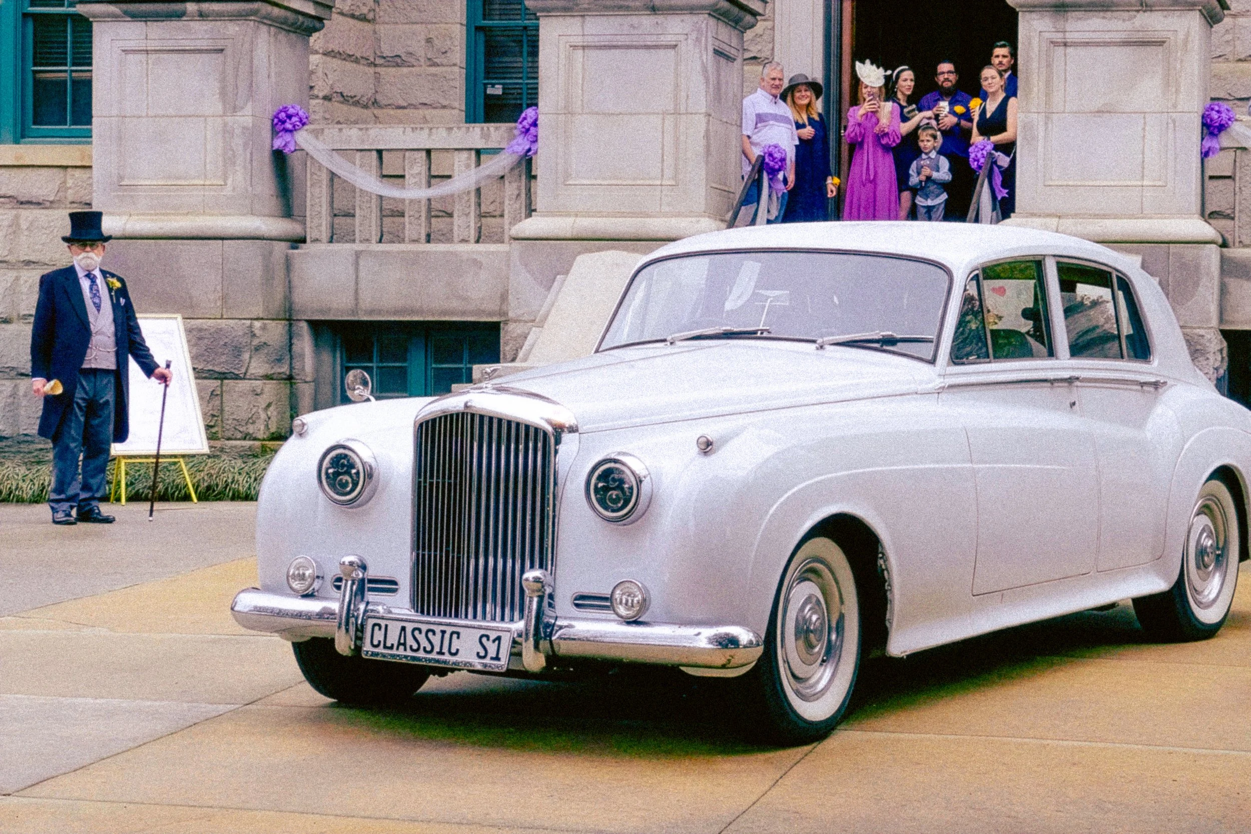 A vintage white classic car parked outside a stone building with people gathered at the entrance, some holding drinks, including women in elegant dresses and hats, and an older man in a suit and top hat, suggesting a celebratory event.