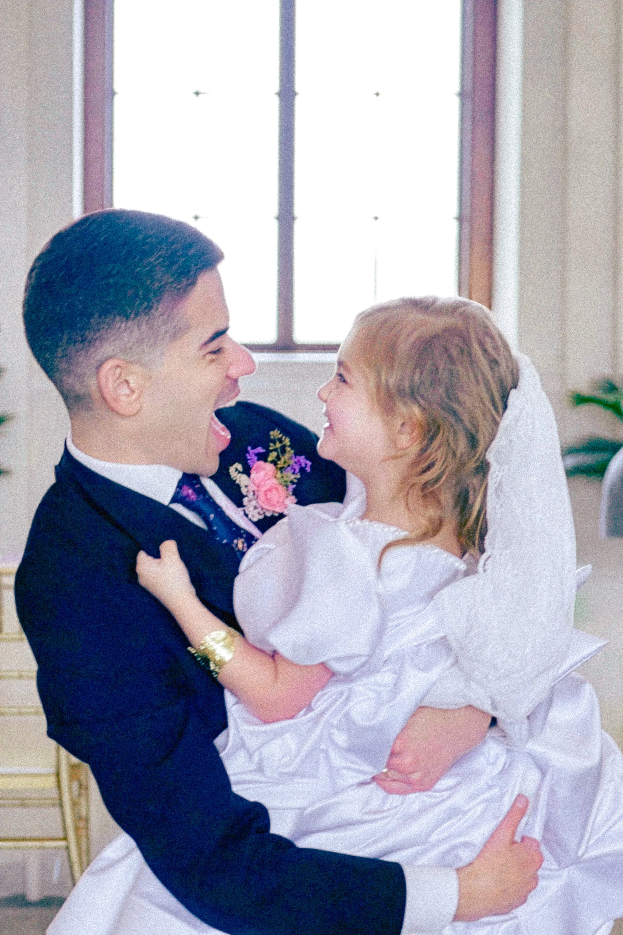 A couple dancing closely together in formal attire, the man wearing a tuxedo with a floral boutonniere, the woman in a white dress, both smiling and looking at each other with a large window in the background.