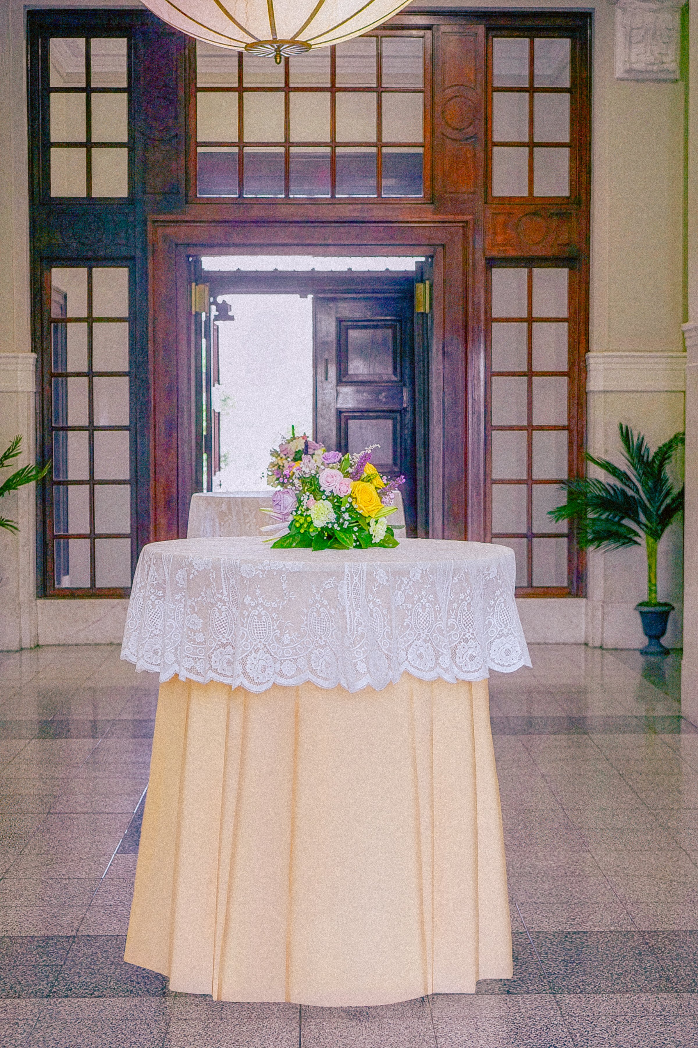 Round table covered with a lace tablecloth and decorated with a colorful flower arrangement, set in a room with wooden-paneled entrance and potted plants.
