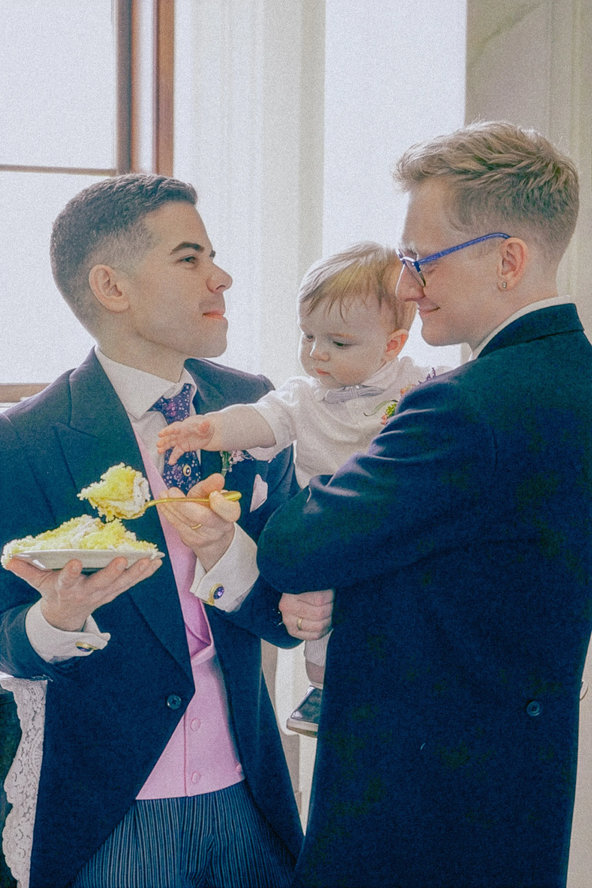 Two men in suits sharing a moment with a young child, who is reaching out to one man. The man on the left is holding a plate with cake, and the child is reaching towards the cake.