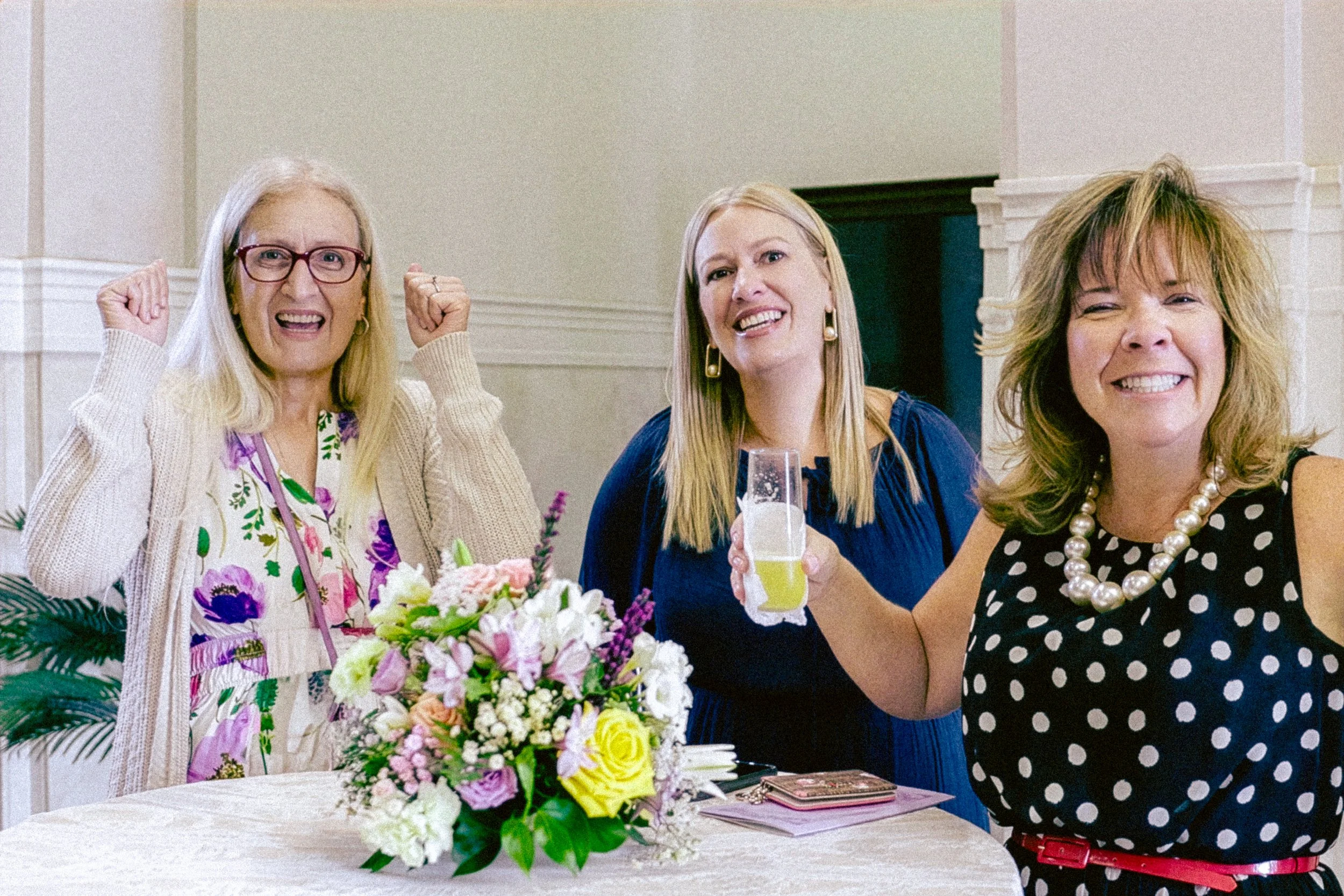 Three women smiling at a celebration, with a floral centerpiece and drinks on the table.