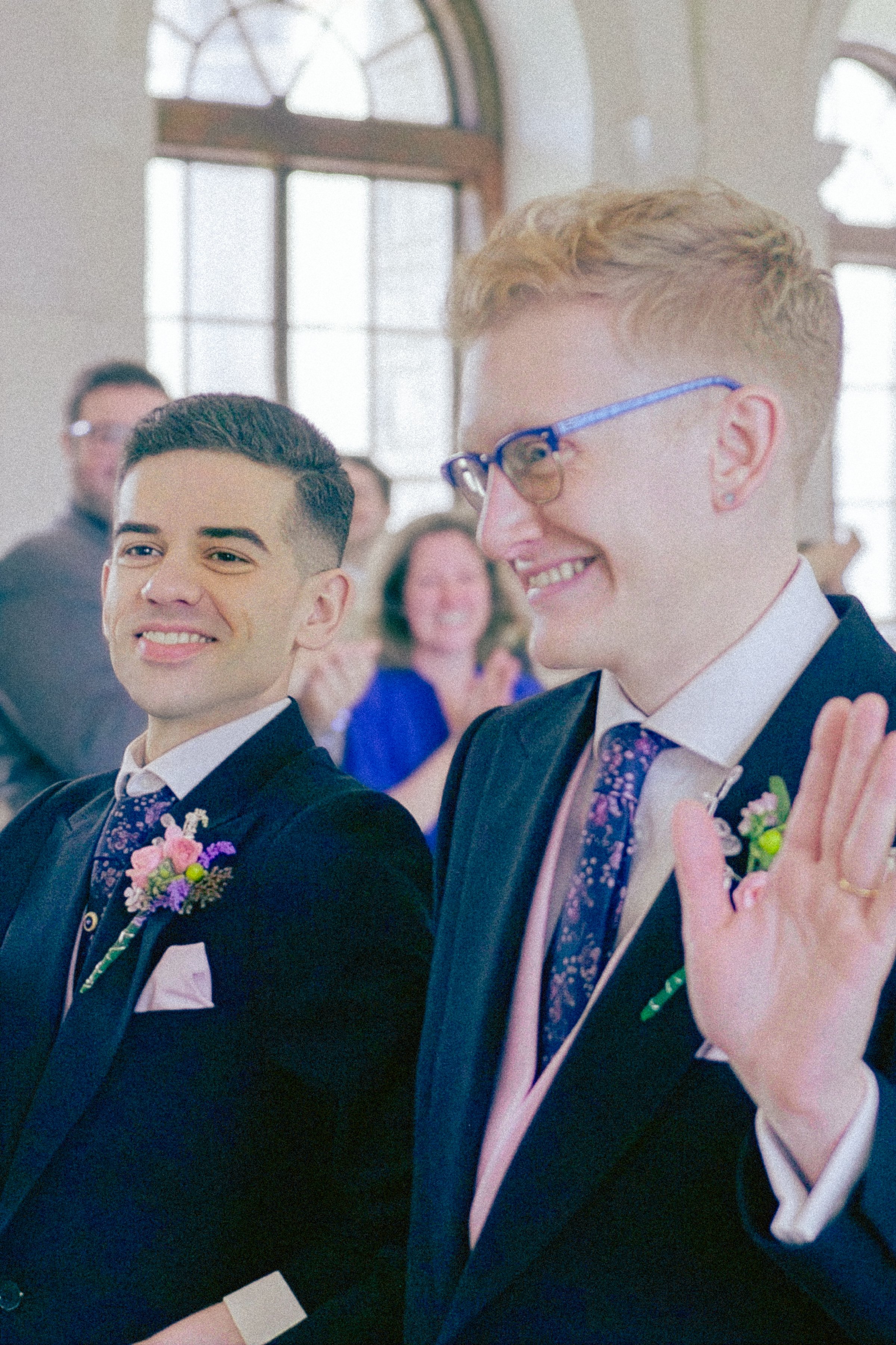 Two young men dressed in formal attire, smiling during a wedding ceremony, with a woman in the background clapping and wearing a blue dress.