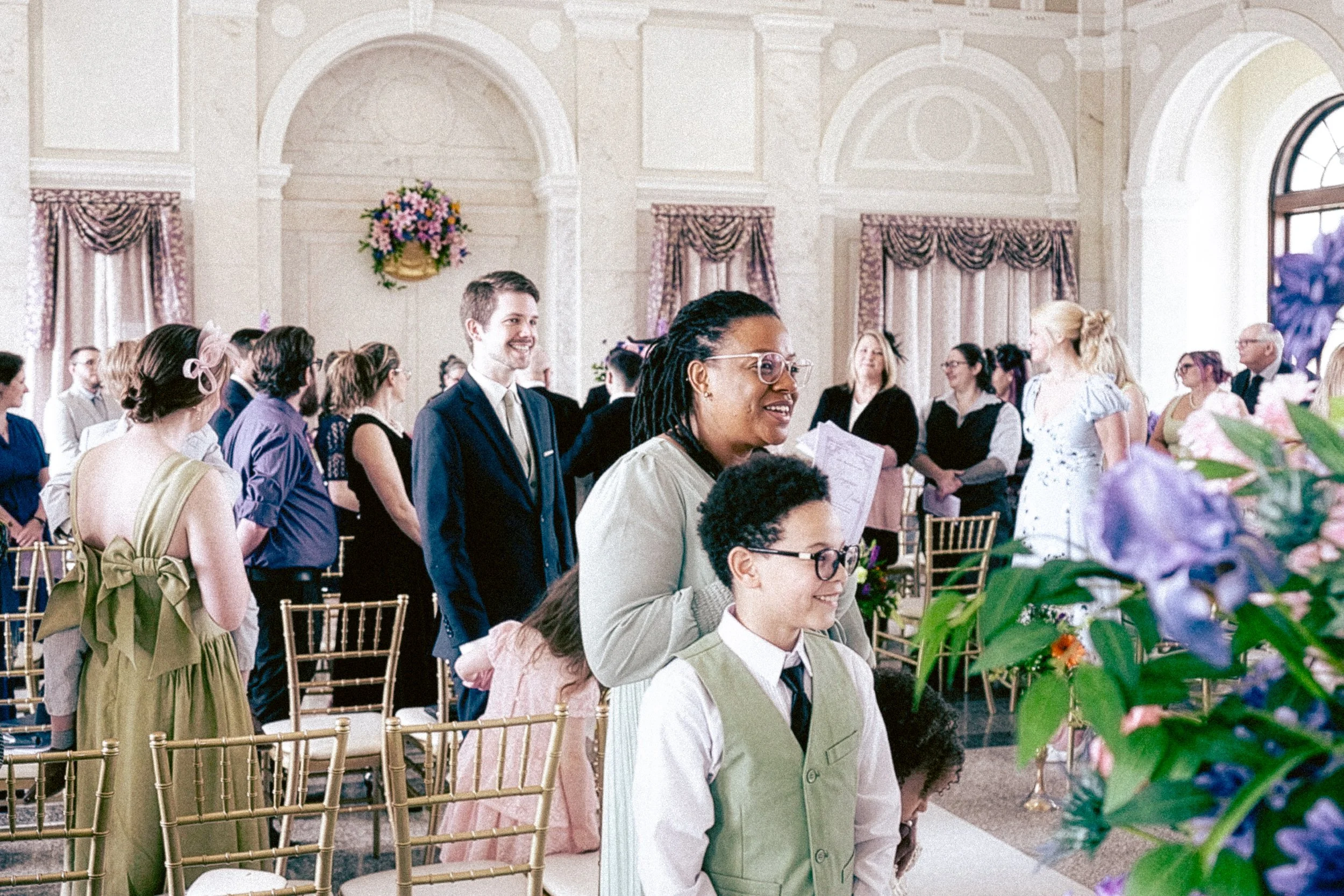 People standing and seated at a wedding ceremony in an elegant, decorated hall with large windows and floral arrangements.
