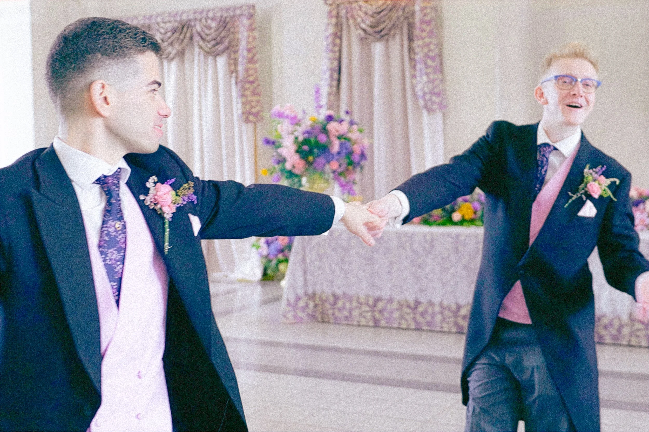 Two men dressed in suits holding hands and dancing at a wedding reception with floral arrangements and ornate curtains in the background.
