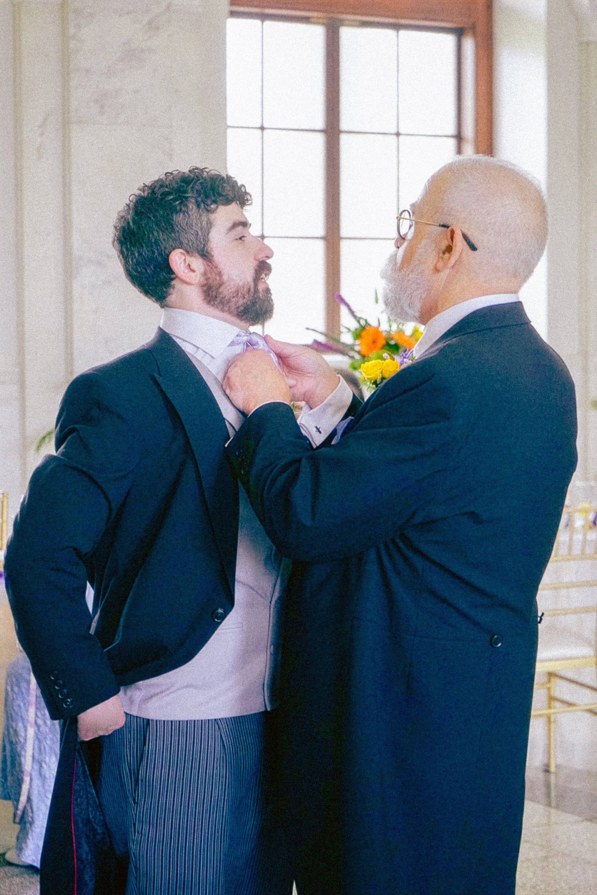 An older man with glasses adjusting the collar of a younger man with a beard. Both are dressed in suits and are standing close together indoors with a large window and flowers in the background.
