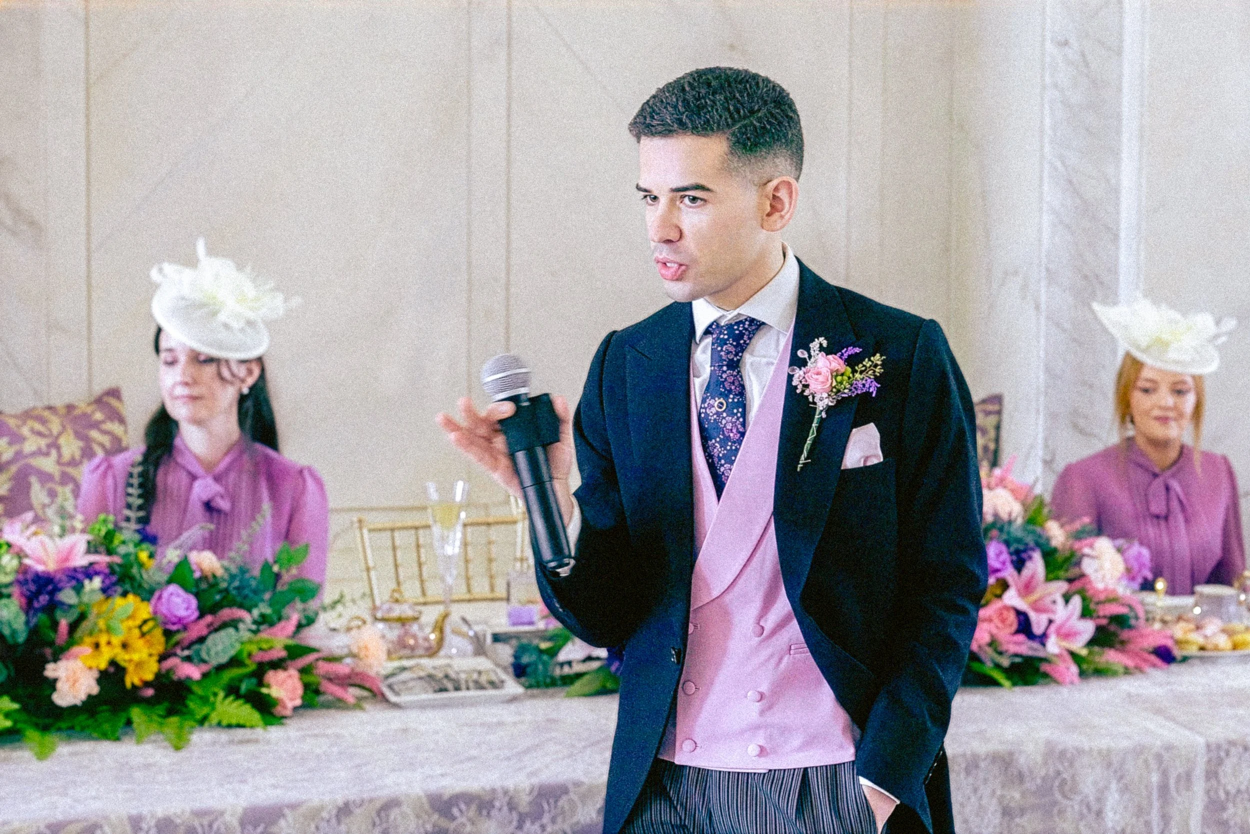 A man in a dark suit and pink vest giving a speech at a wedding reception, holding a microphone, with women in purple dresses wearing fancy hats and floral arrangements on the table in the background.
