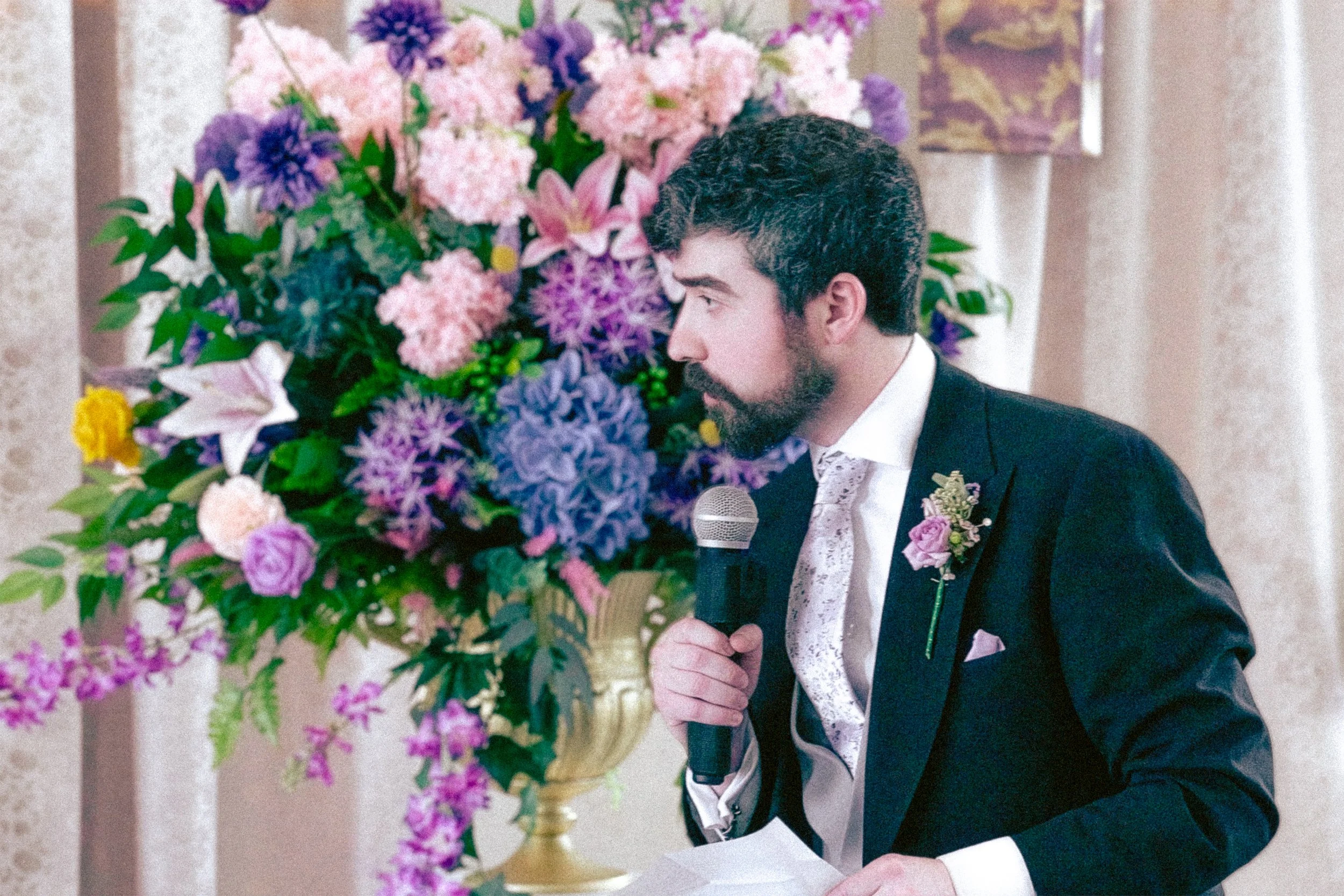 A man in a tuxedo with a boutonniere is speaking into a microphone near a large bouquet of pink, purple, and white flowers at an indoor event.