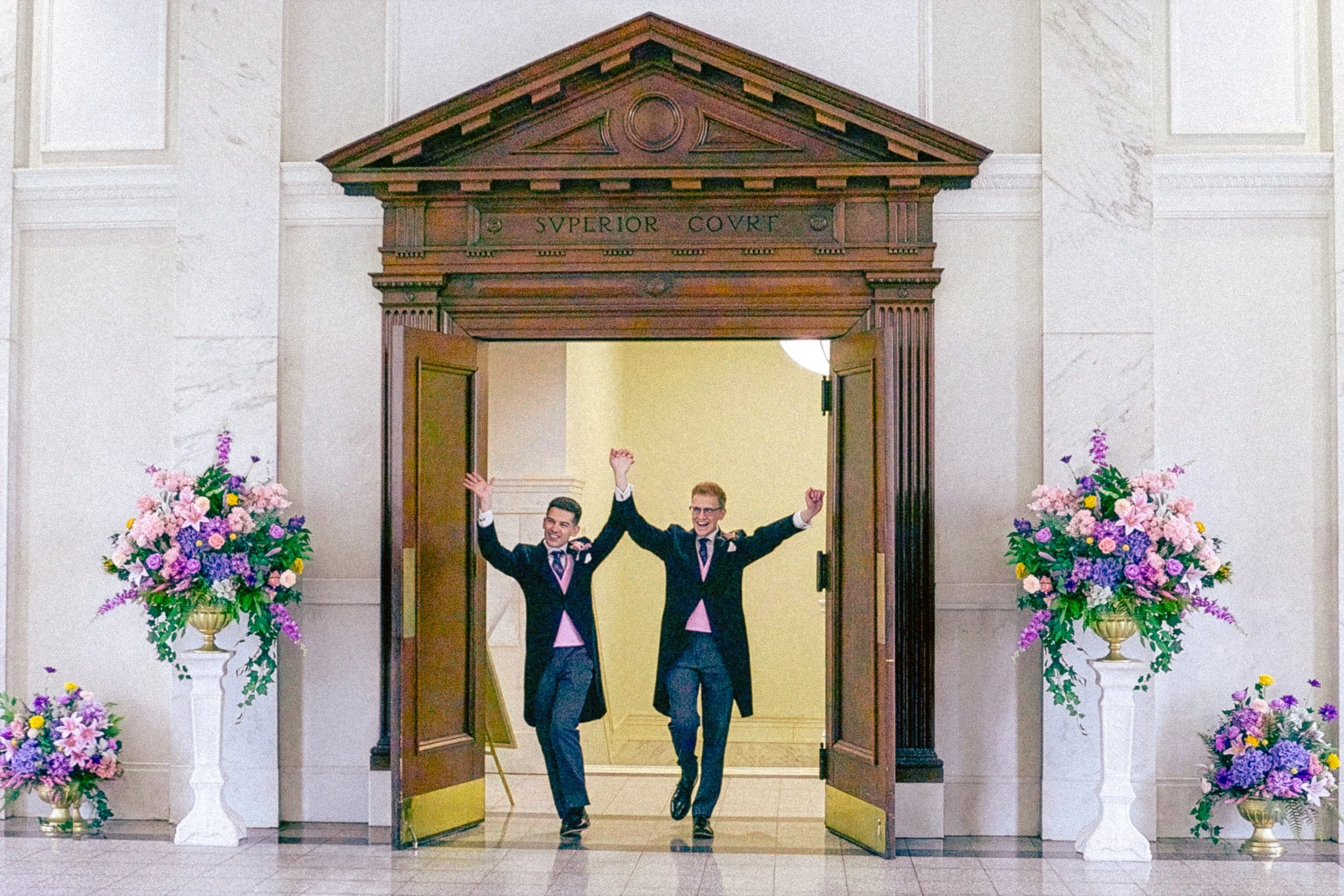 Two men in tuxedos celebrating in a doorway inside a building, surrounded by large flower arrangements.