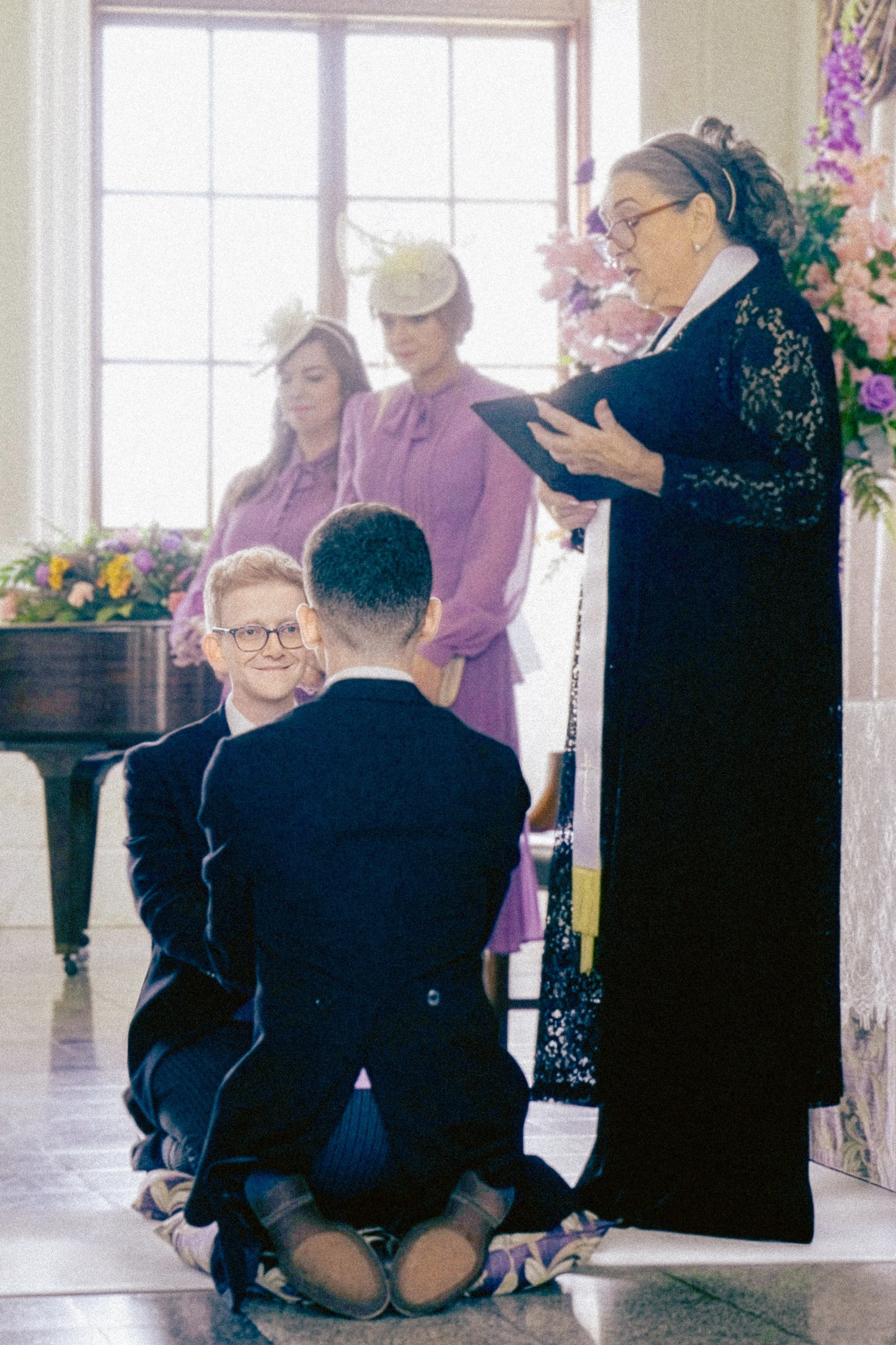 A woman with glasses reading from a black book or folder while standing in front of an audience in a room decorated with flowers. Two children sit on the floor in front of her, facing her with their backs to the camera. The children appear to be prep