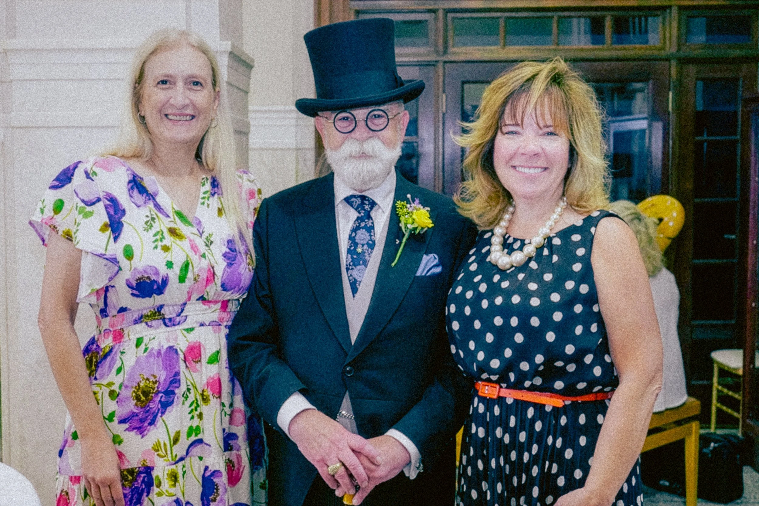 Three smiling people, two women and one man, standing together indoors. The man in the middle is wearing a top hat, glasses, and a suit with a boutonniere, and the women are dressed in colorful, patterned dresses. All are posing for a photo, with the