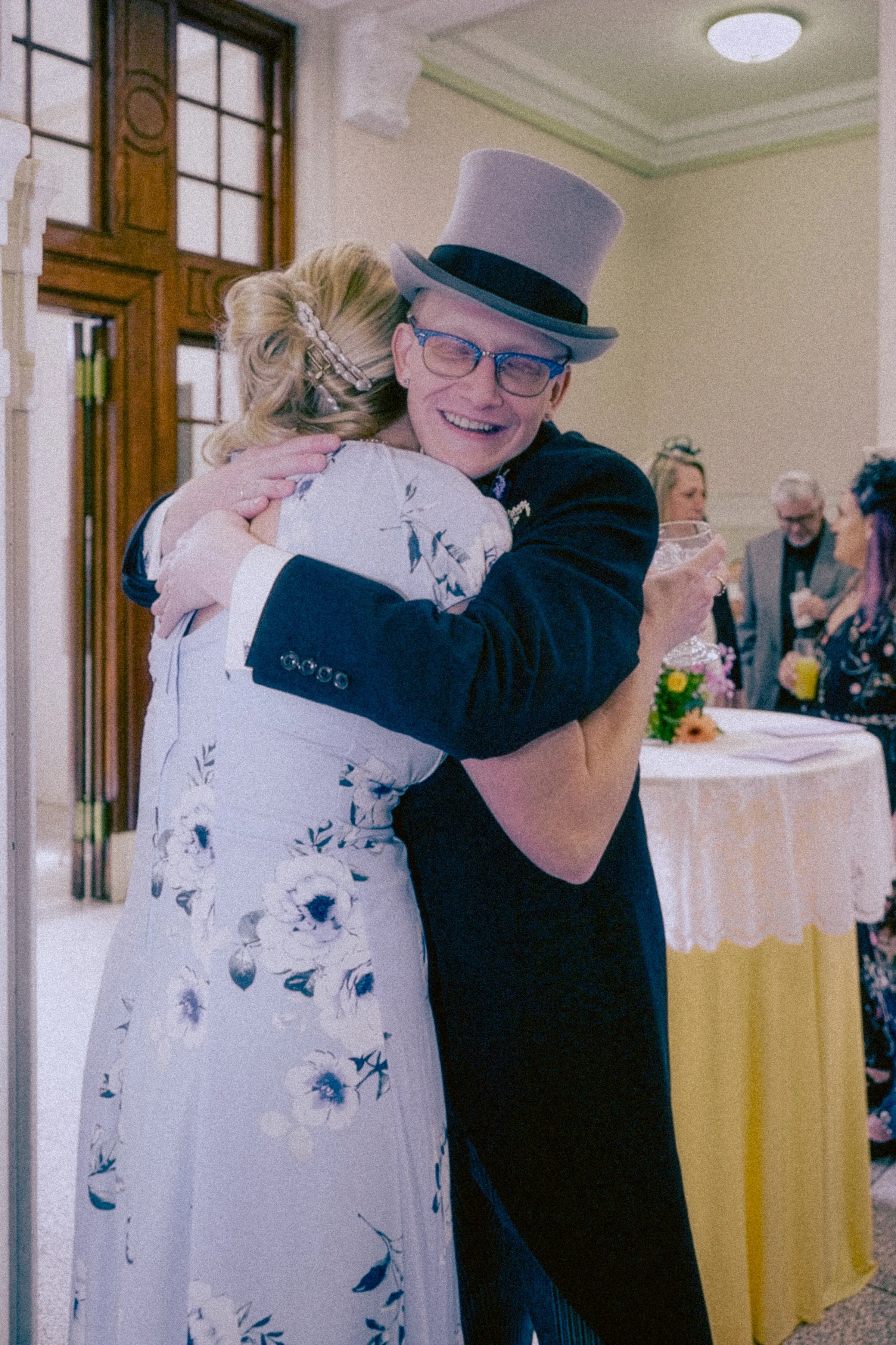 A man wearing a top hat, glasses, and a dark suit hugging a woman in a floral dress at a celebration or formal event.