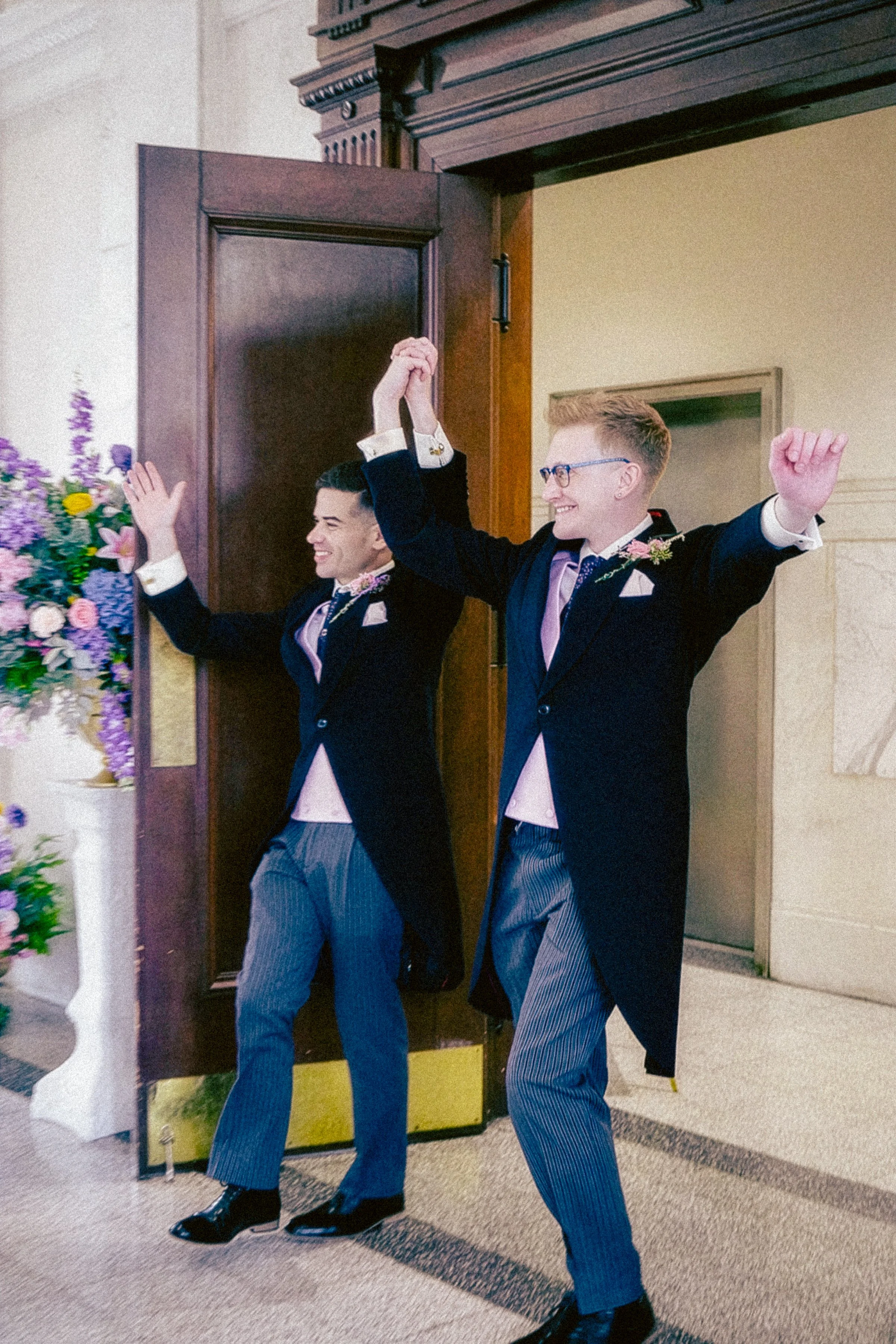 Two men dressed in formal suits are celebrating inside a building, with one man holding their hands together above their heads and the other smiling with arms raised. They appear happy and are wearing striped trousers and bow ties. There is a floral 