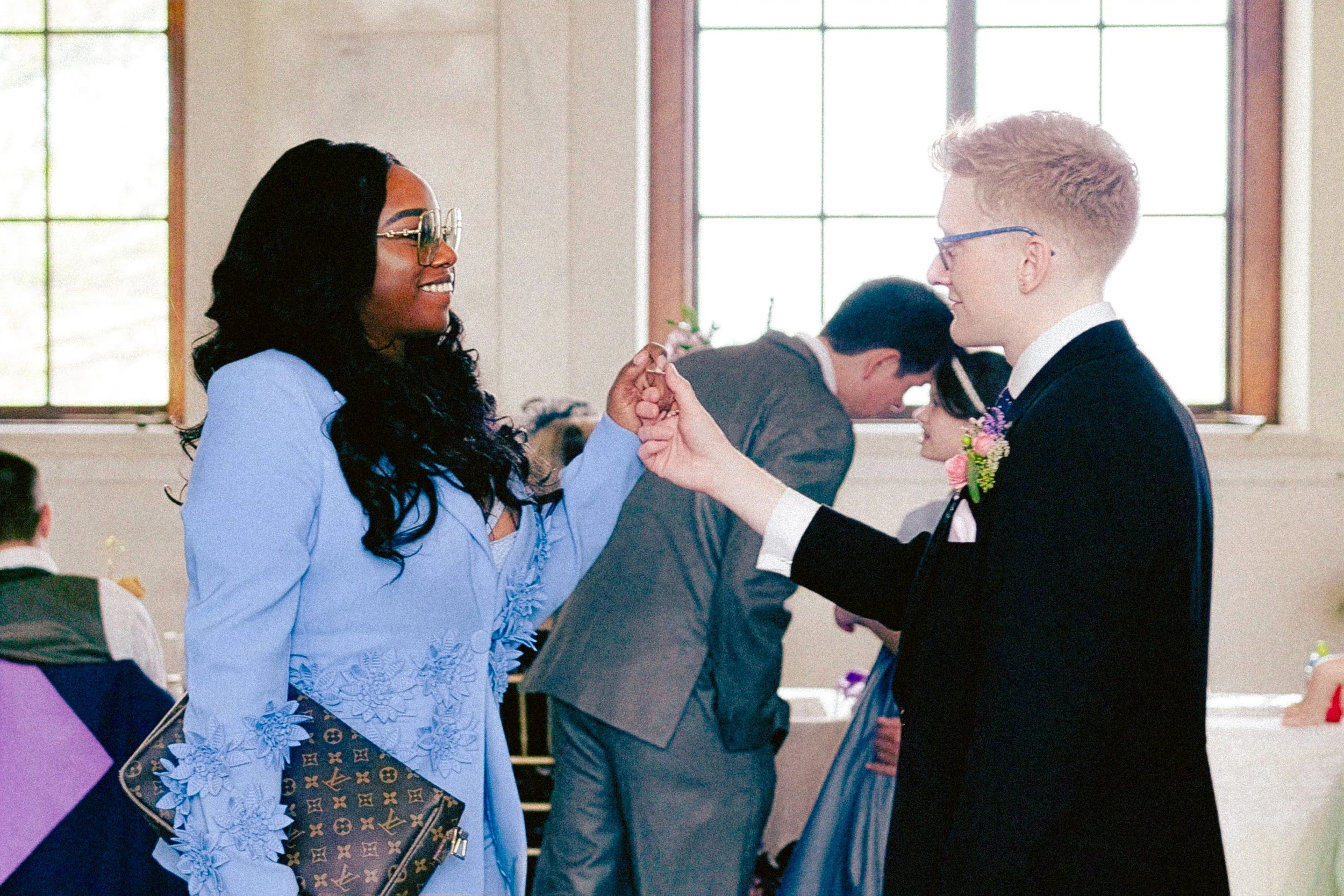 A woman in a light blue dress with glasses and a handbag holding hands with a man in a suit and glasses, both smiling, inside a room with large windows, possibly at a formal event.