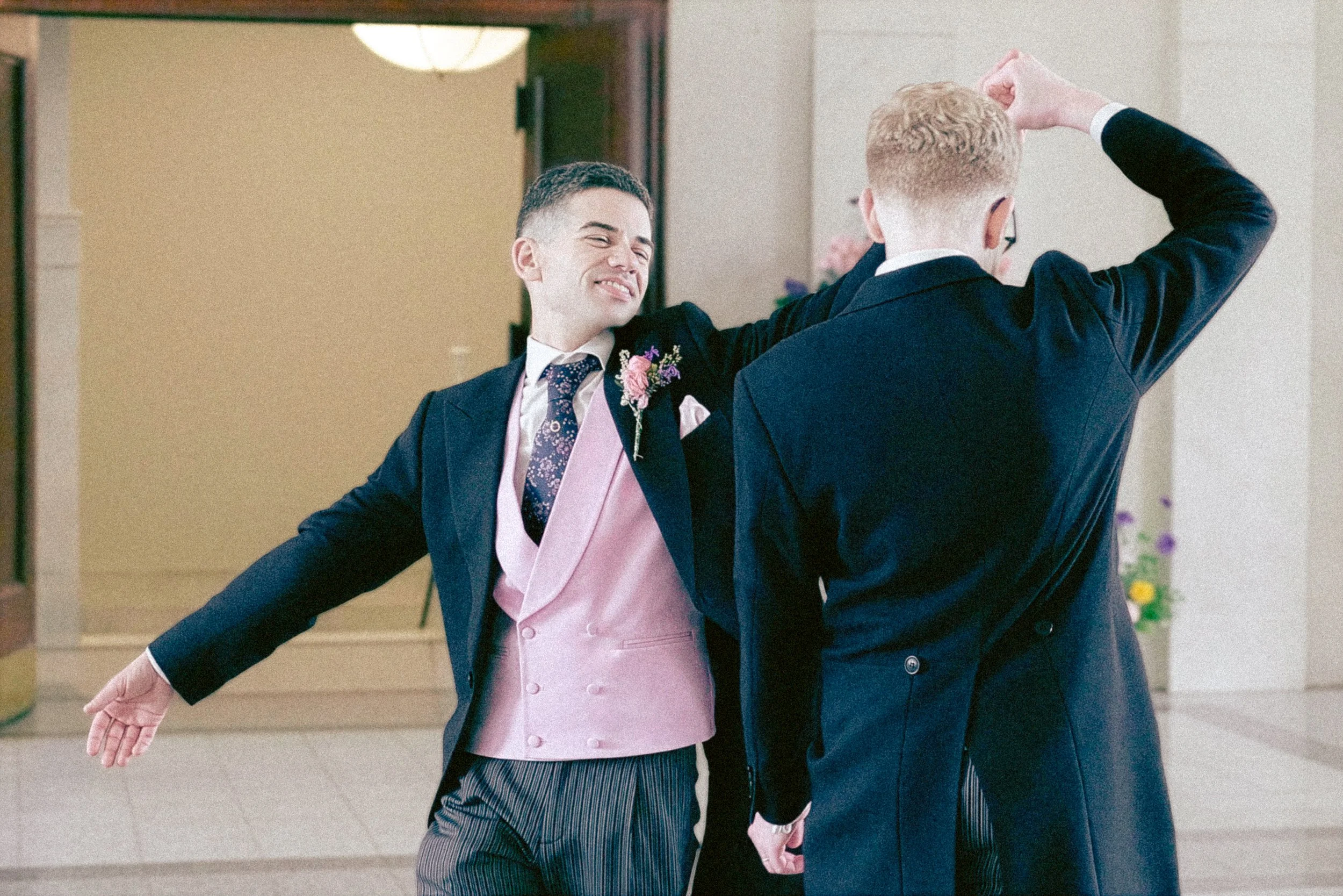 Two men in formal attire dancing indoors, one with arms outstretched and smiling, the other with back to camera raising one hand, with a decorated background.