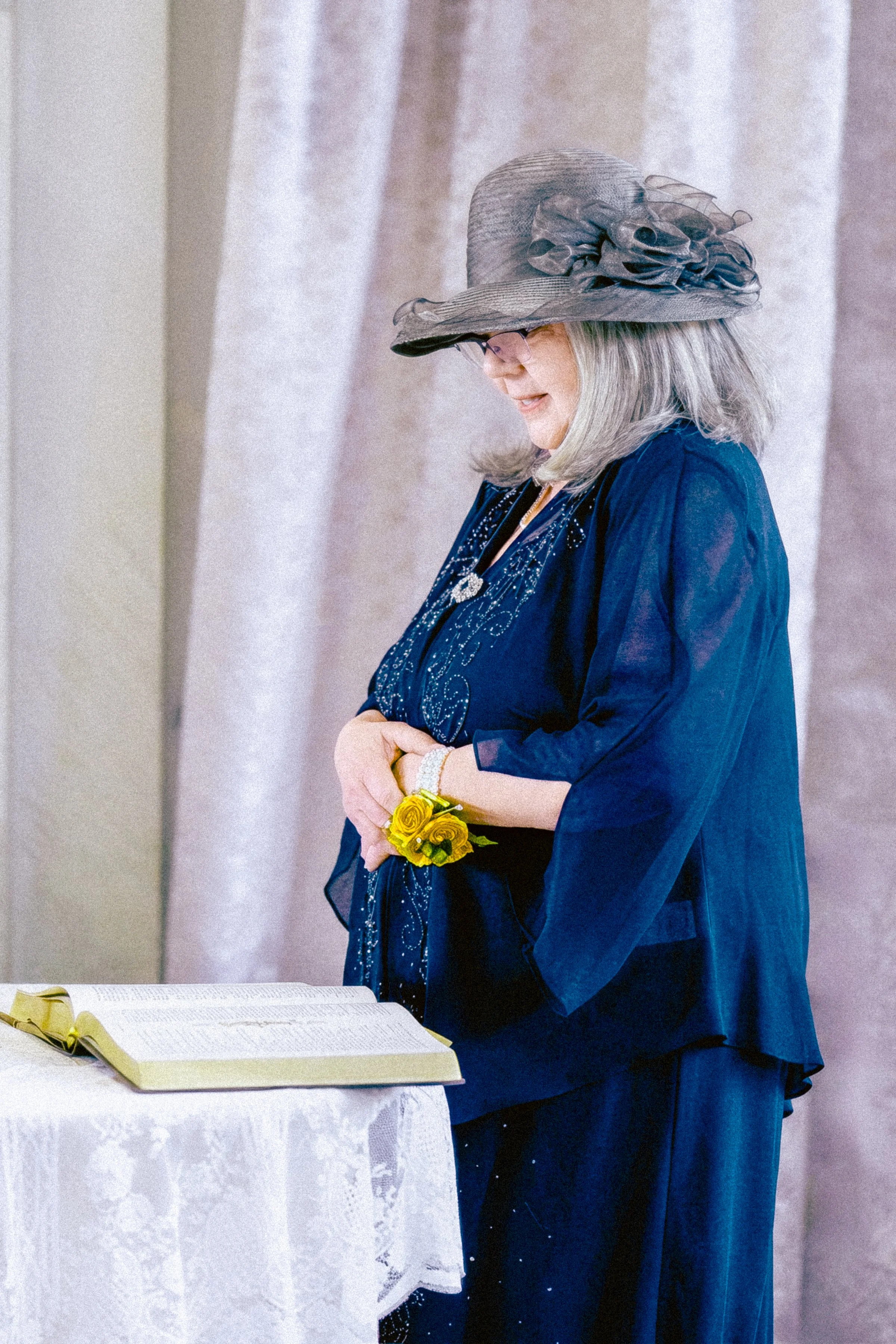 A woman with gray hair wearing a large gray hat with a ruffle adornment, a dark blue embroidered top, and black pants with shiny embellishments, holding yellow roses and standing near an open book on a white tablecloth.