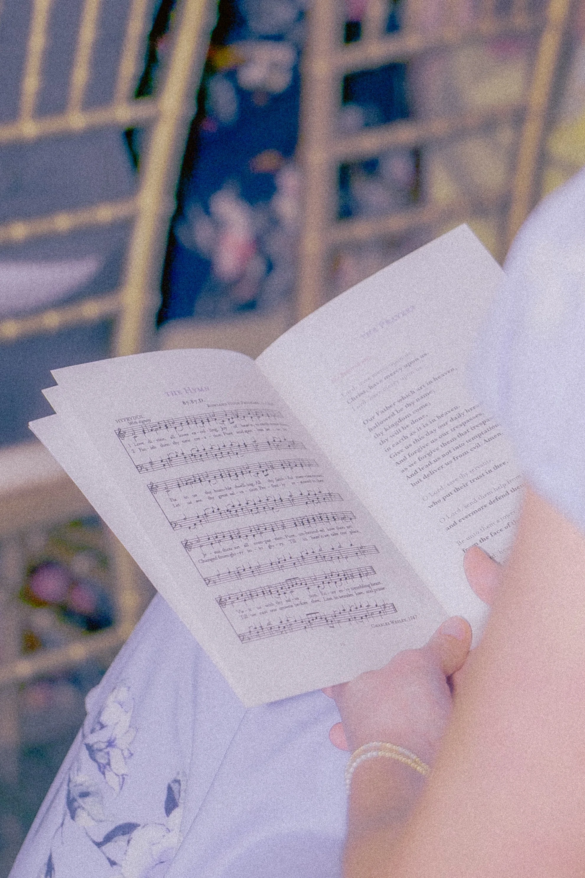 A person is holding a pamphlet open to a page with sheet music and text, with a background of a building with barred windows and some greenery.