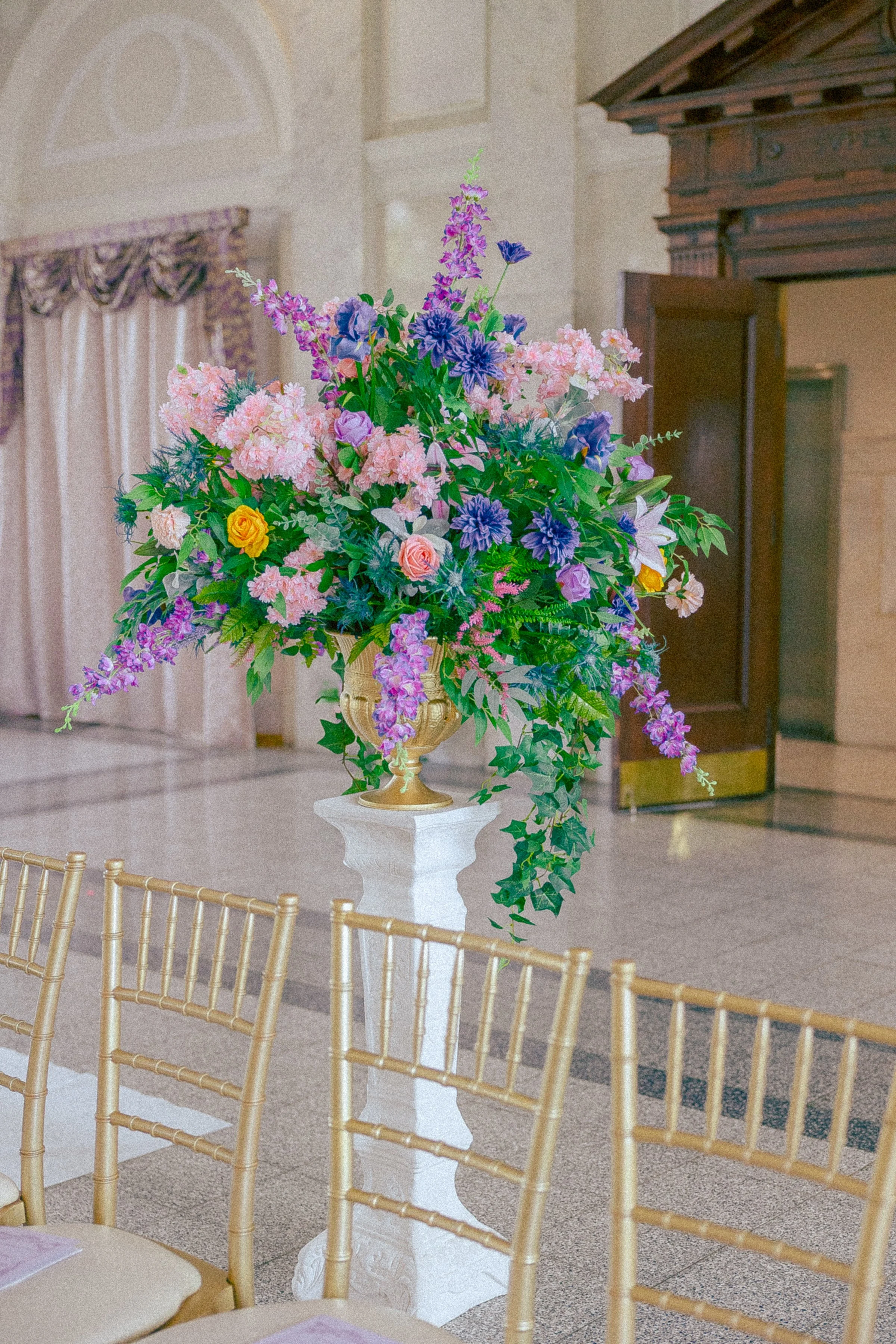 Flower arrangement with pink, purple, yellow, and white flowers on a white pedestal in a decorated event hall.