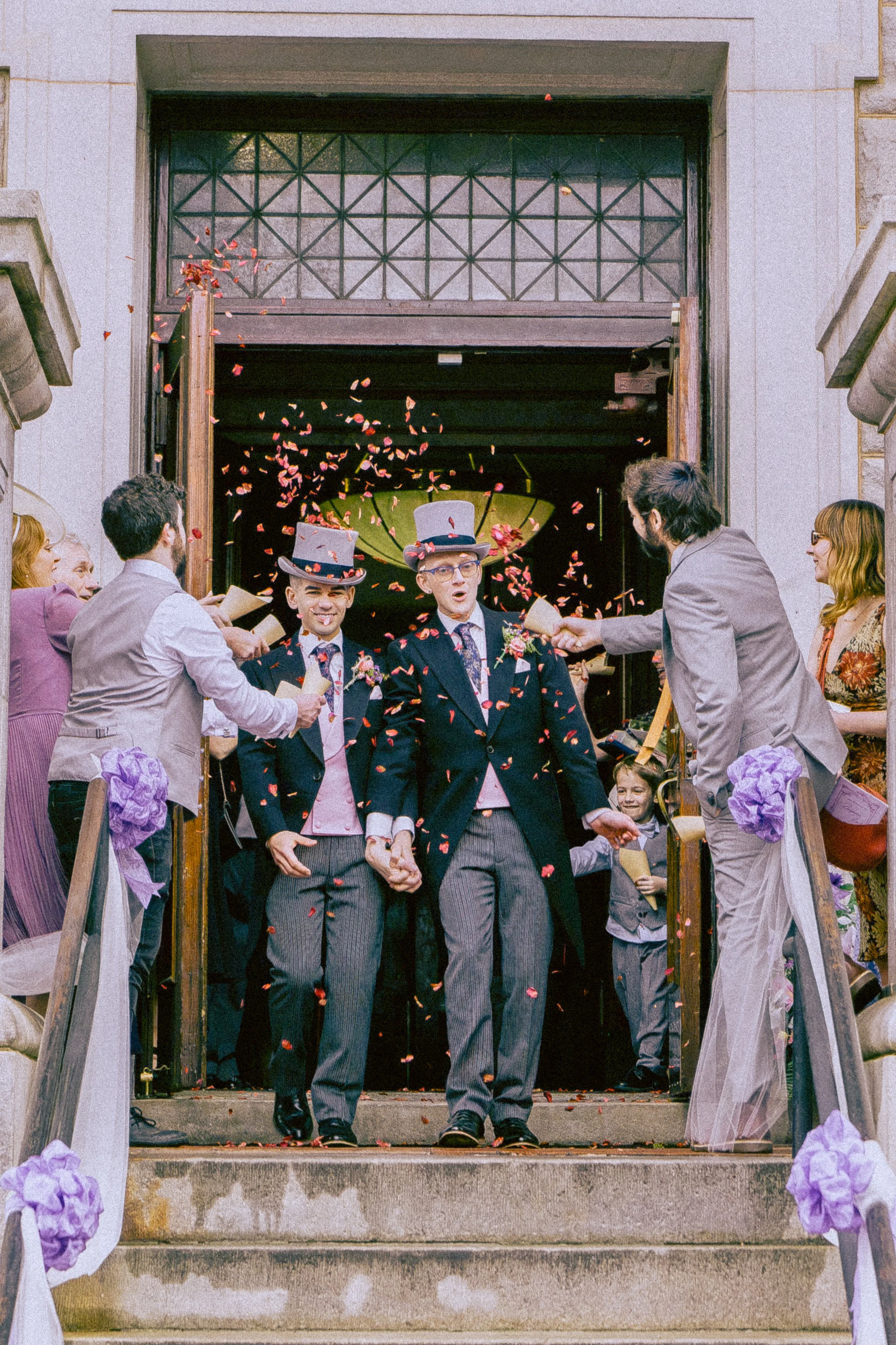 Two men in tuxedos holding hands and walking down the steps as a wedding celebration with guests and family cheering and throwing flower petals.